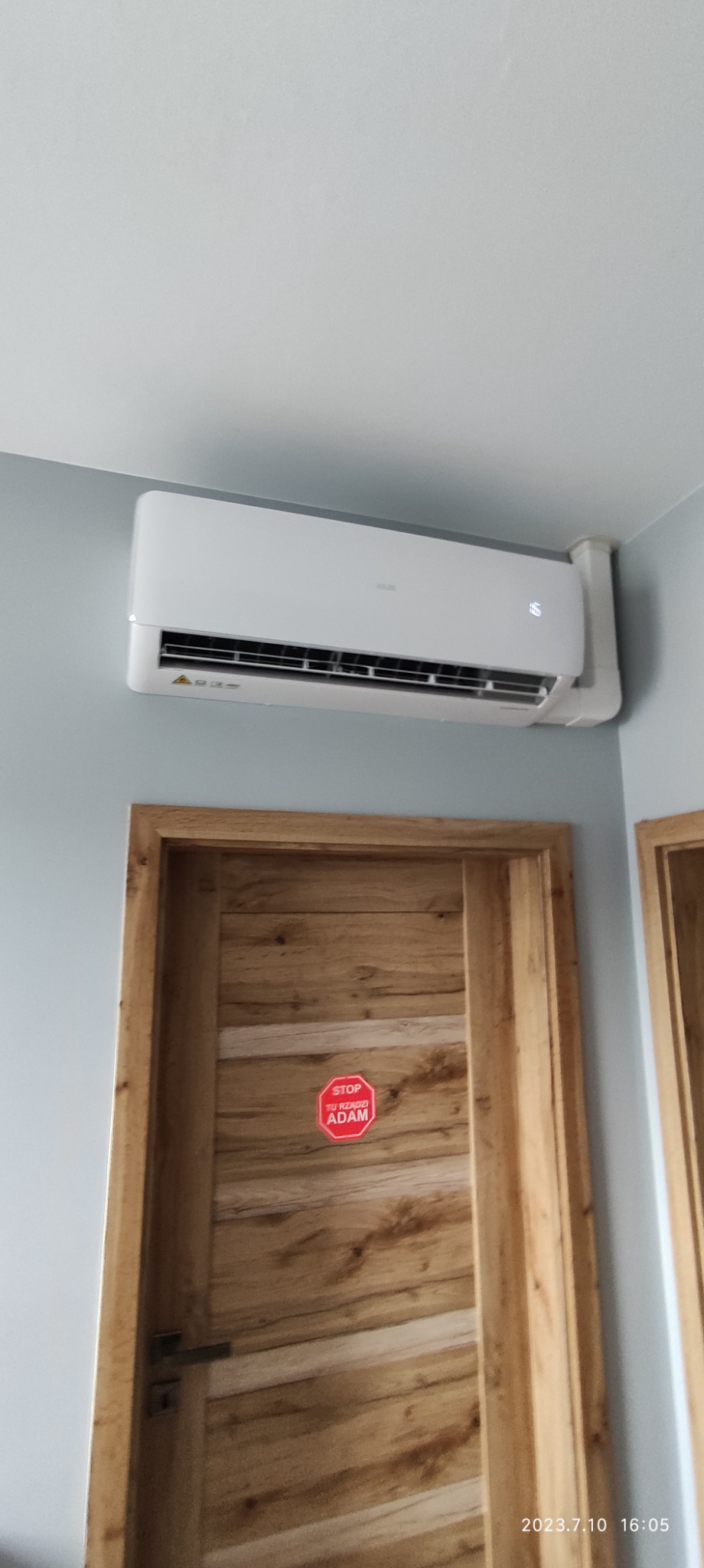 Biały klimatyzator ścienny zamontowany nad drewnianymi drzwiami z naklejką 'STOP Tu Rządzi ADAM'.