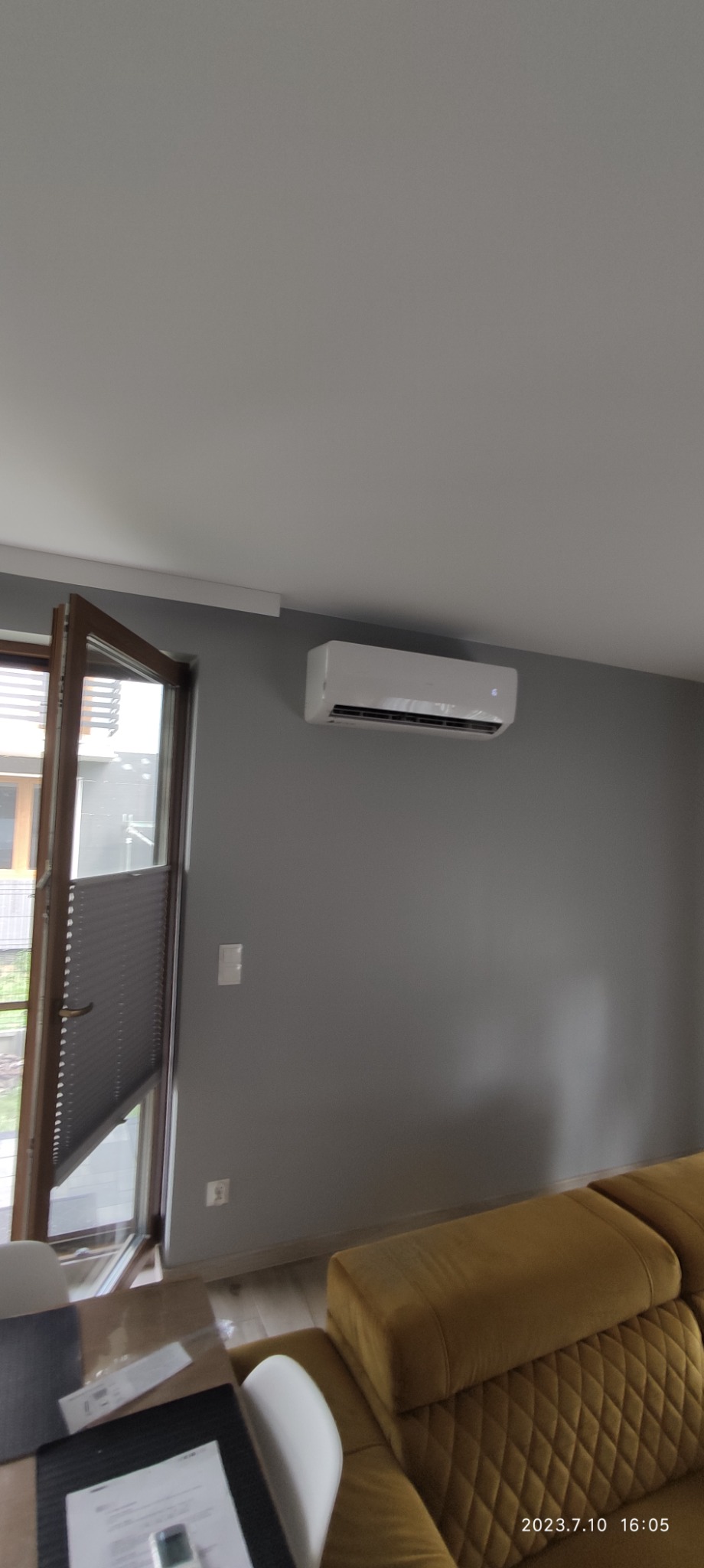 Biała klimatyzacja ścienna zamontowana w szarym pokoju z otwartym oknem i żaluzjami plisowanymi, widoczny fragment żółtej sofy.
