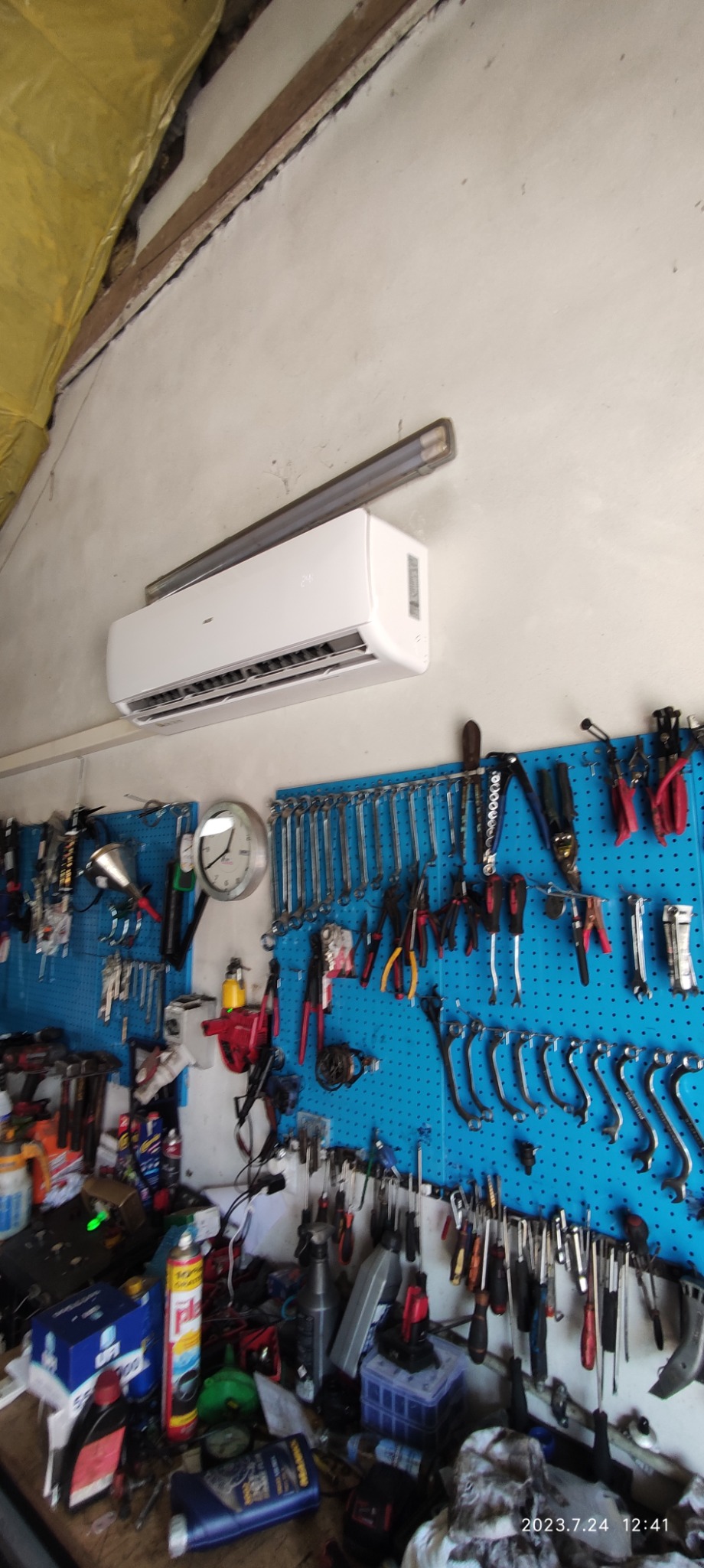 Biały klimatyzator ścienny zamontowany w warsztacie, z niebieską tablicą na narzędzia i różnymi kluczami oraz narzędziami wiszącymi pod spodem.