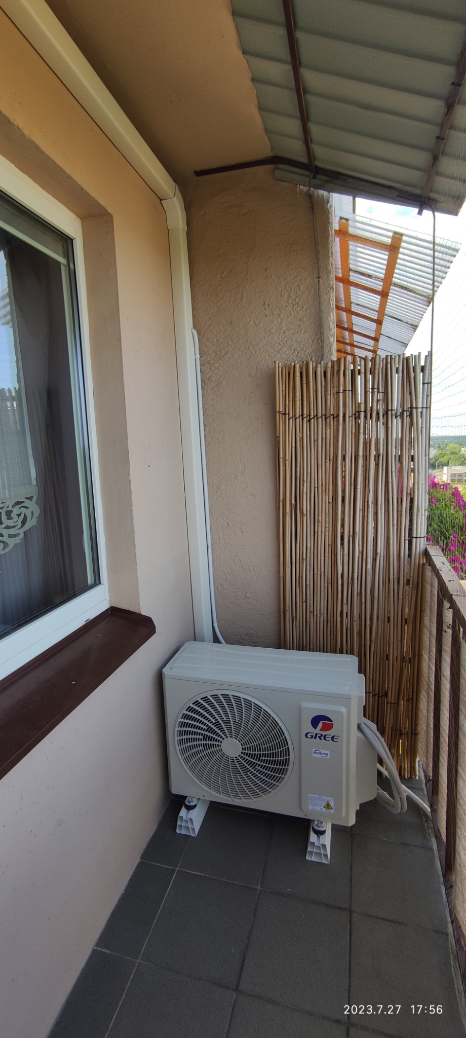 Biała jednostka zewnętrzna klimatyzatora marki Gree zamontowana na balkonie, widoczna ściana budynku z oknem i bambusowa osłona balkonu.