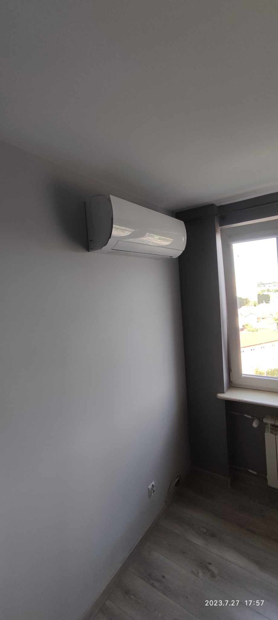 Biały klimatyzator ścienny zamontowany w szarym pokoju, widok z dołu, okno z widokiem na miasto w tle.