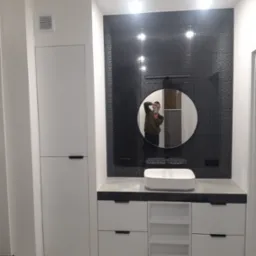 Nowoczesna łazienka z białą szafką pod umywalką, okrągłym lustrem i czarną mozaiką na ścianie, widoczne odbicie osoby robiącej zdjęcie.