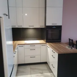 Nowoczesna kuchnia z białymi, błyszczącymi frontami szafek, czarnymi uchwytami i drewnianym blatem, oświetlona złotą lampą wiszącą, z widoczną piekarnikiem w zabudowie i marmurową podłogą.