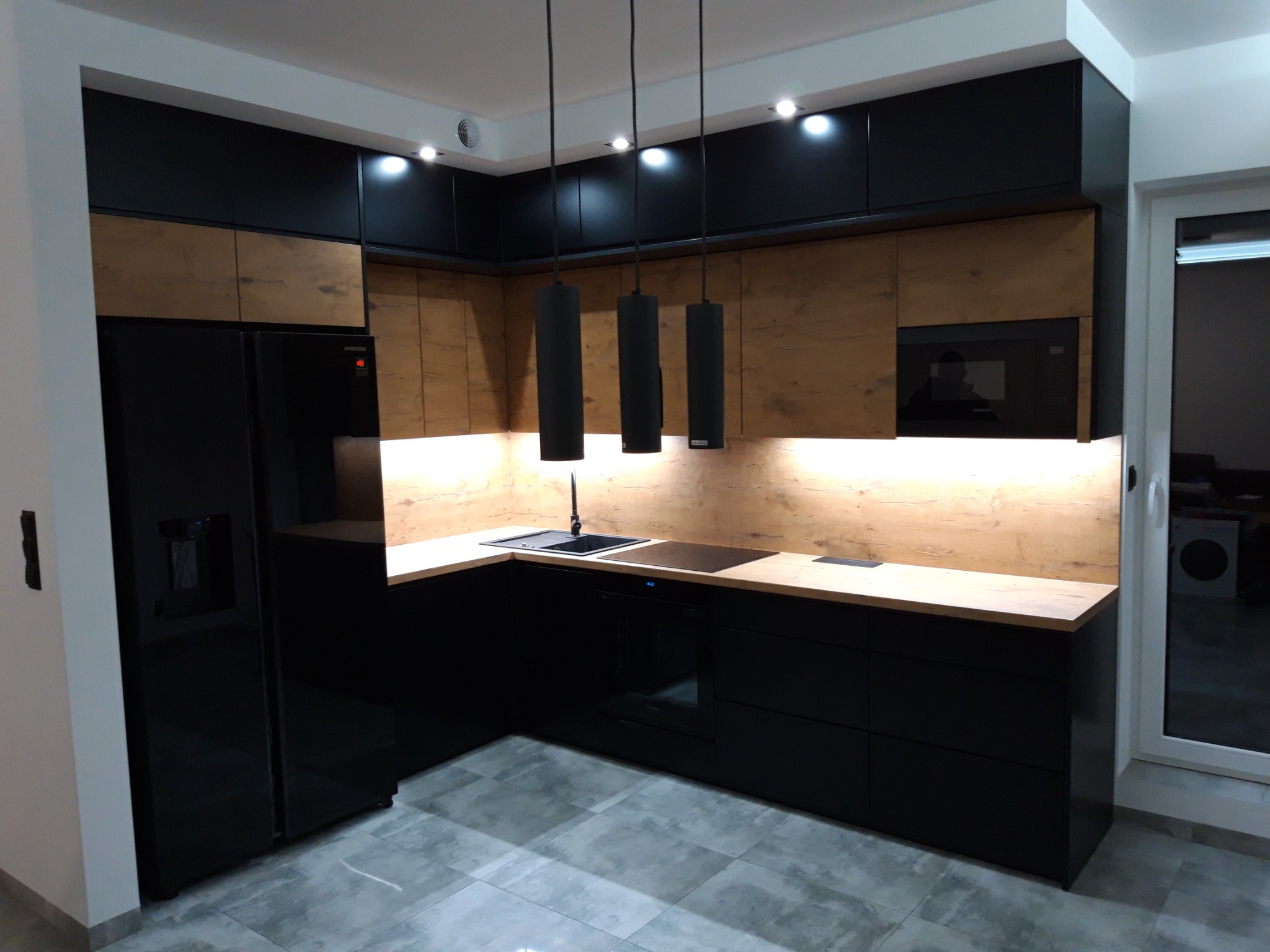 Nowoczesna kuchnia z czarnymi szafkami i drewnianymi panelami, oświetlona wiszącymi lampami cylindrycznymi, z blatem w odcieniu drewna i szarą podłogą.