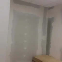 Ściana w trakcie szpachlowania z widocznymi niedoskonałościami i przygotowanymi płytami gipsowymi w rogu pokoju, oświetlenie sufitowe.