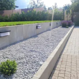 Oświetlenie LED zamontowane na betonowym murku wzdłuż ścieżki z białego żwiru, z zielenią w tle i brukowaną ścieżką z boku.