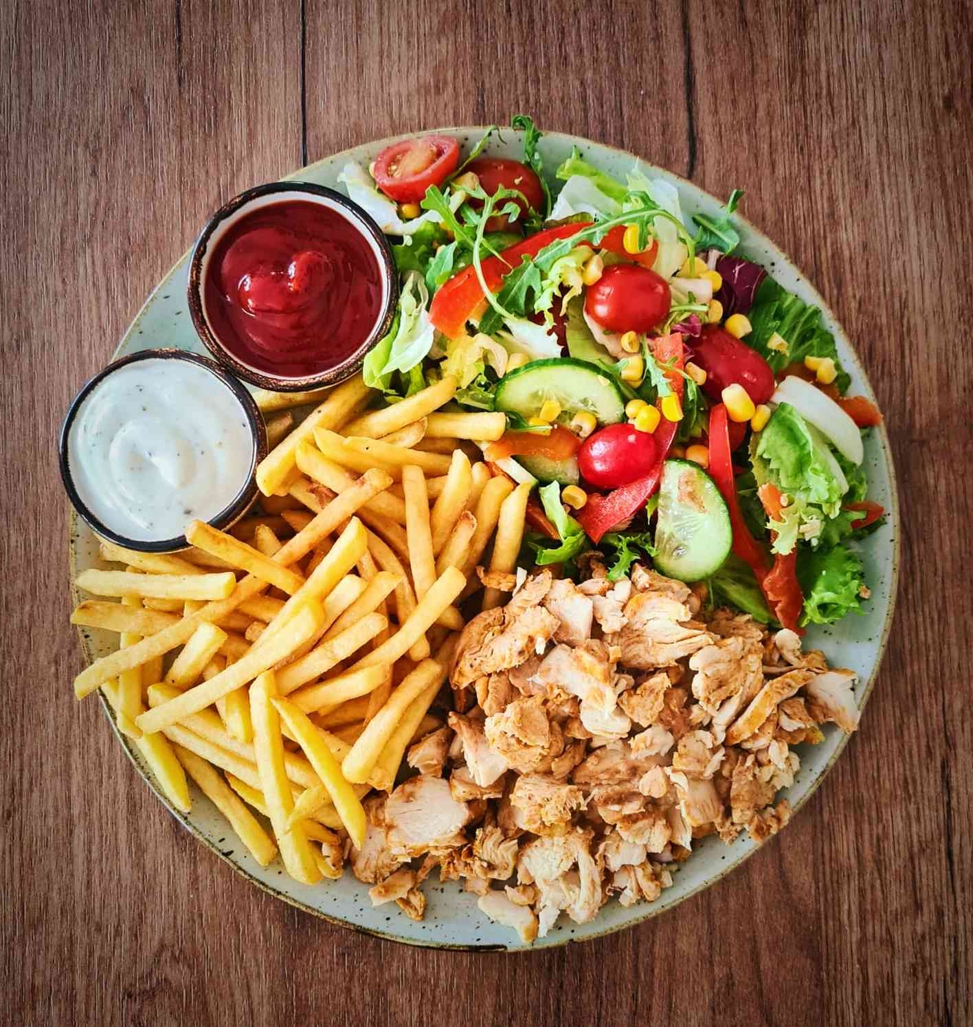 Porcja kebaba drobiowego na talerzu z frytkami, sałatką warzywną i dwoma sosami, całość ułożona na drewnianym stole.