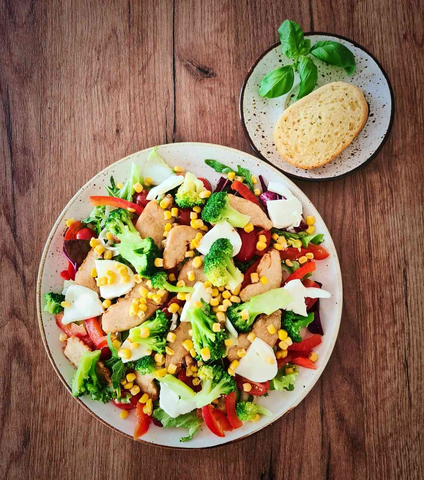 Sałatka z pieczoną piersią kurczaka, brokułami, kukurydzą, mozzarellą, papryką i pomidorami koktajlowymi na białym talerzu z brązową obwódką, obok talerzyk z kromką chleba i bazylią, na drewnianym...