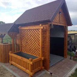 Drewniana konstrukcja z zadaszeniem z brązowej dach&oacute;wki, treliażem i donicą, stylizowana na altanę z elementami warsztatu w tle.