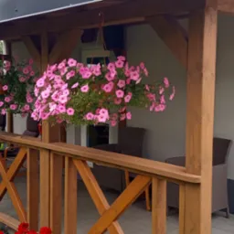 Zadaszony taras z drewnianą balustradą, wiszące donice z różowymi kwiatami, wiklinowe krzesła w tle.