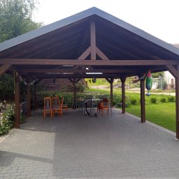 Jelonek Janusz Gil - Drewniana konstrukcja z zadaszeniem na posesji, z meblami ogrodowymi, donicami i kostką brukową na podłożu. Widoczny skuter i dekoracje.