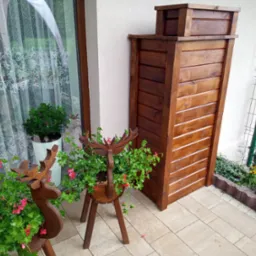 Brązowy pojemnik na śmieci z drewna obok okna i drewnianych kwietników w kształcie jeleni z kwitnącymi pelargoniami na wyłożonym kostką podwórku.