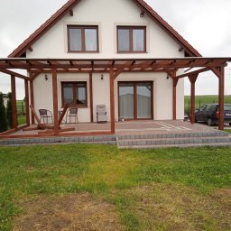 Jelonek Janusz Gil - Drewniana konstrukcja pergoli zadaszająca taras przed domem z białymi ścianami i brązowymi oknami, widoczne krzesła ogrodowe i trawnik.