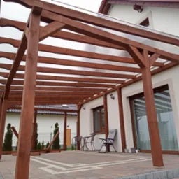 Zadaszona, drewniana konstrukcja z przezroczystym dachem, przylegająca do jasnego domu z widocznymi oknami i meblami ogrodowymi na wybrukowanym tarasie.