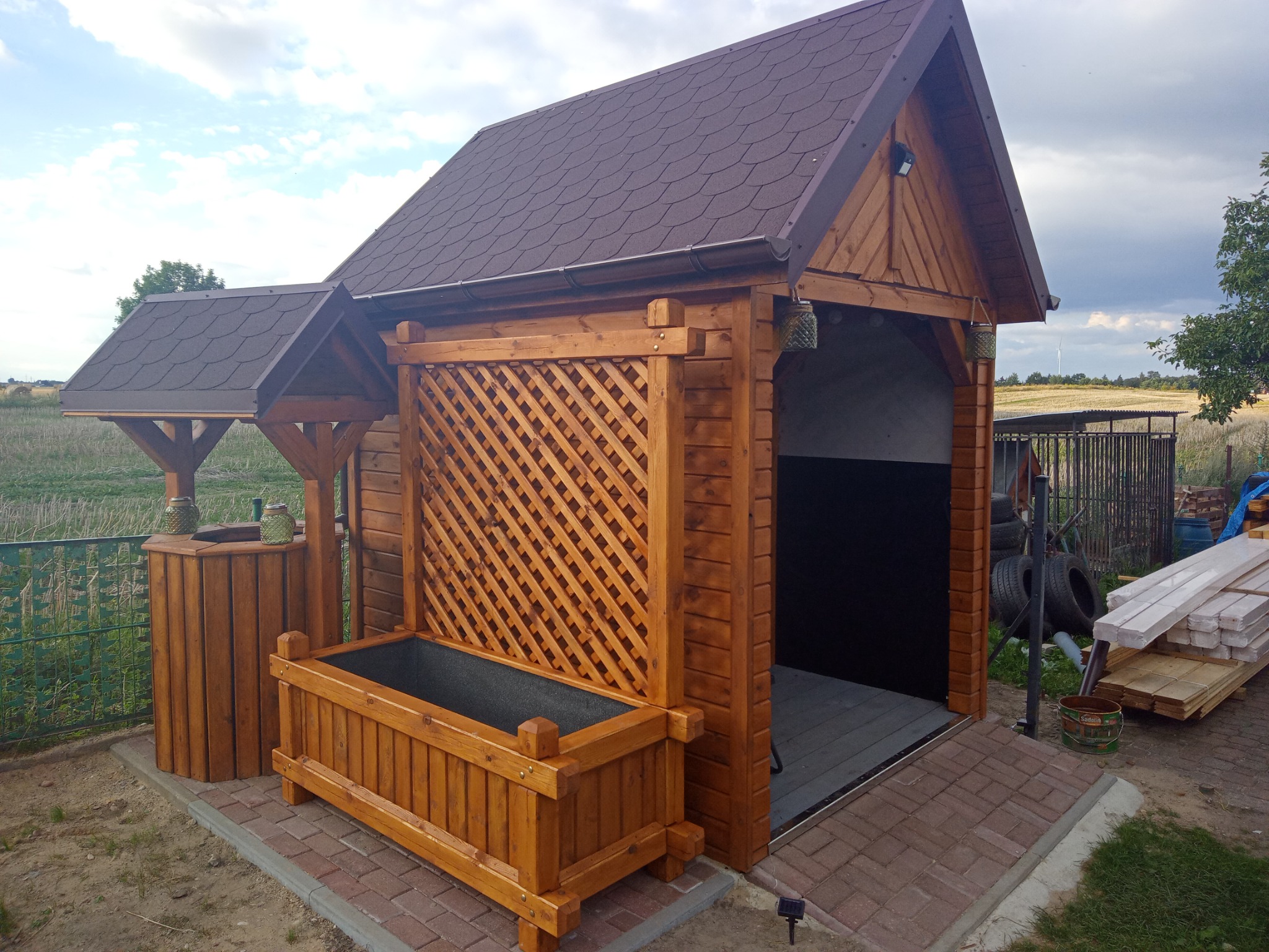 Drewniana konstrukcja z zadaszeniem z brązowej dachówki, treliażem i donicą, stylizowana na altanę z elementami warsztatu w tle.