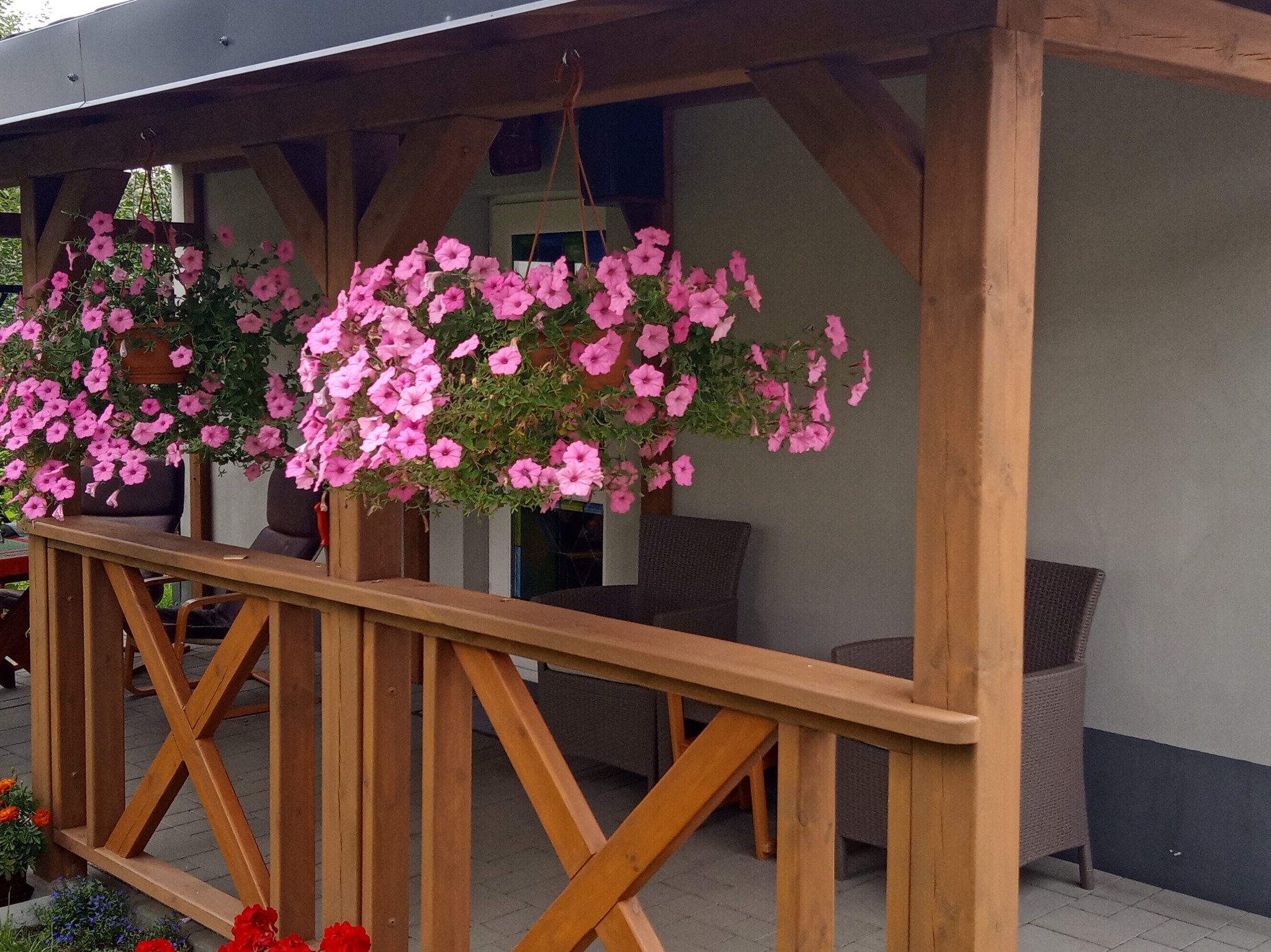 Zadaszony taras z drewnianą balustradą, wiszące donice z różowymi kwiatami, wiklinowe krzesła w tle.