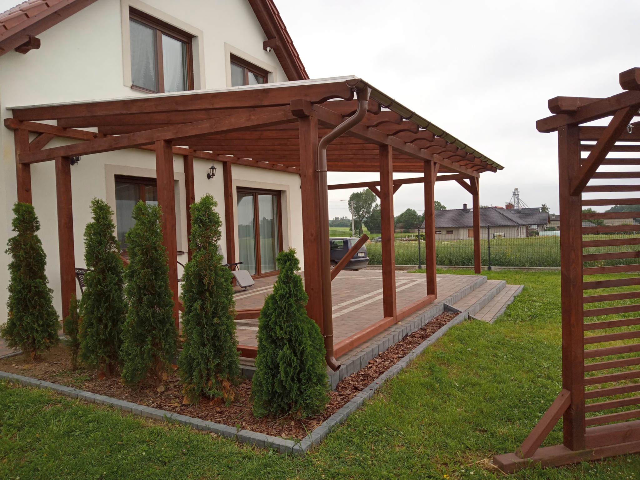 Zadaszony taras przy domu z drewnianą konstrukcją, rynną i młodymi tujami wzdłuż krawędzi, widok na ogród i zabudowania w tle.