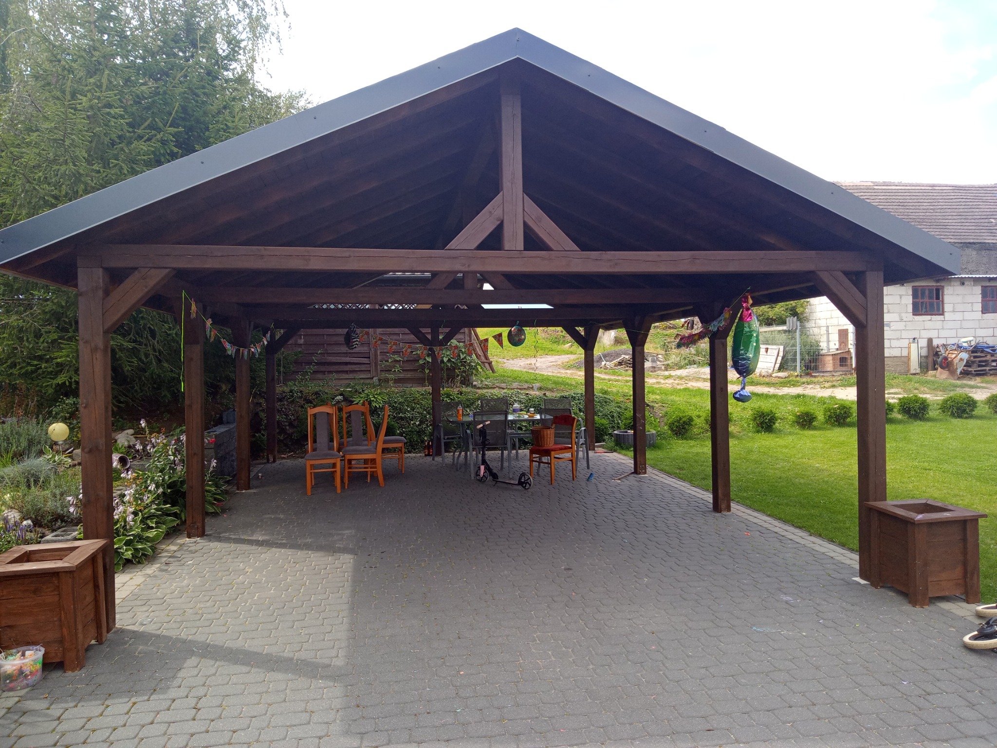 Drewniana konstrukcja z zadaszeniem na posesji, z meblami ogrodowymi, donicami i kostką brukową na podłożu. Widoczny skuter i dekoracje.