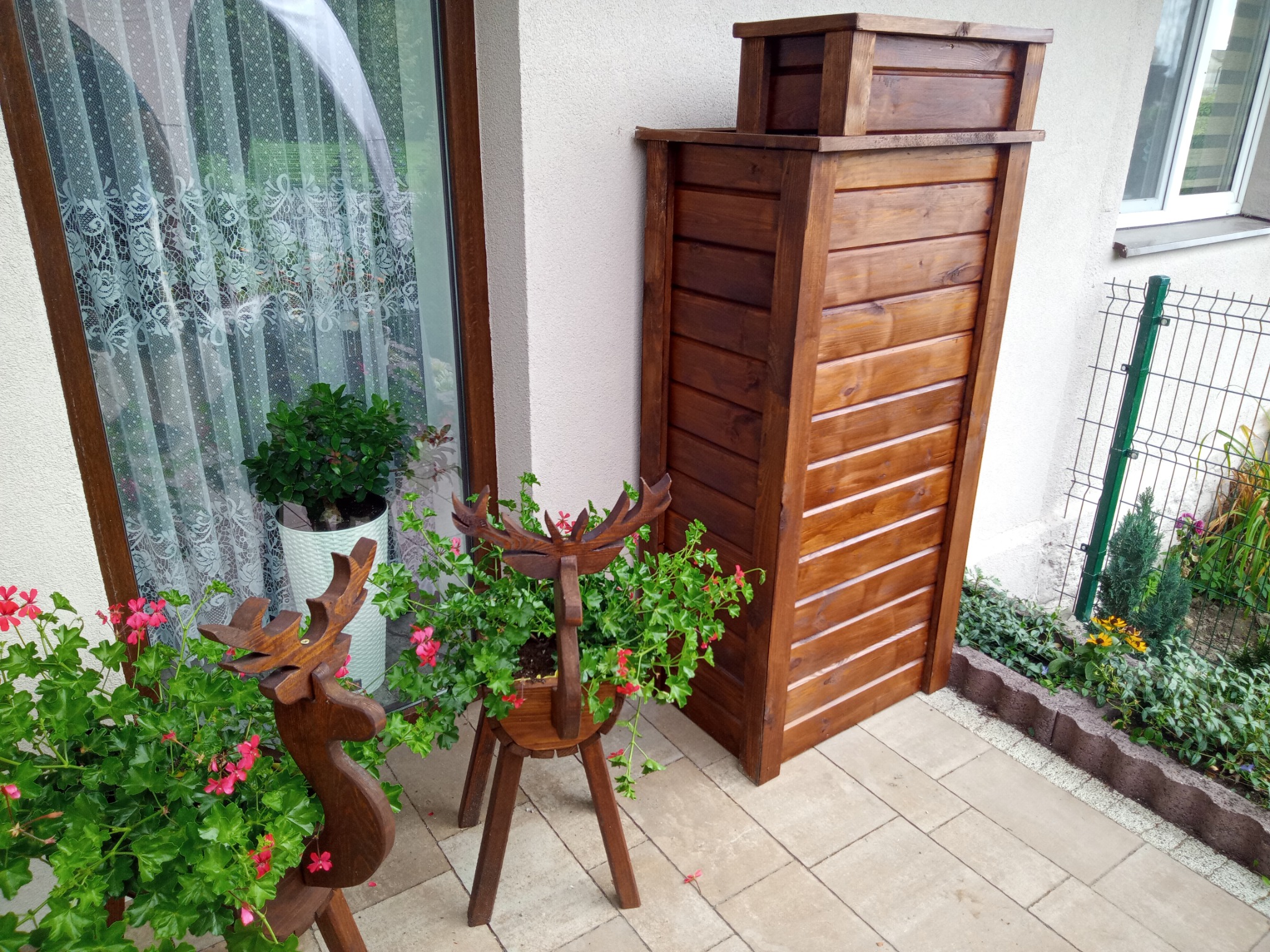 Brązowy pojemnik na śmieci z drewna obok okna i drewnianych kwietników w kształcie jeleni z kwitnącymi pelargoniami na wyłożonym kostką podwórku.