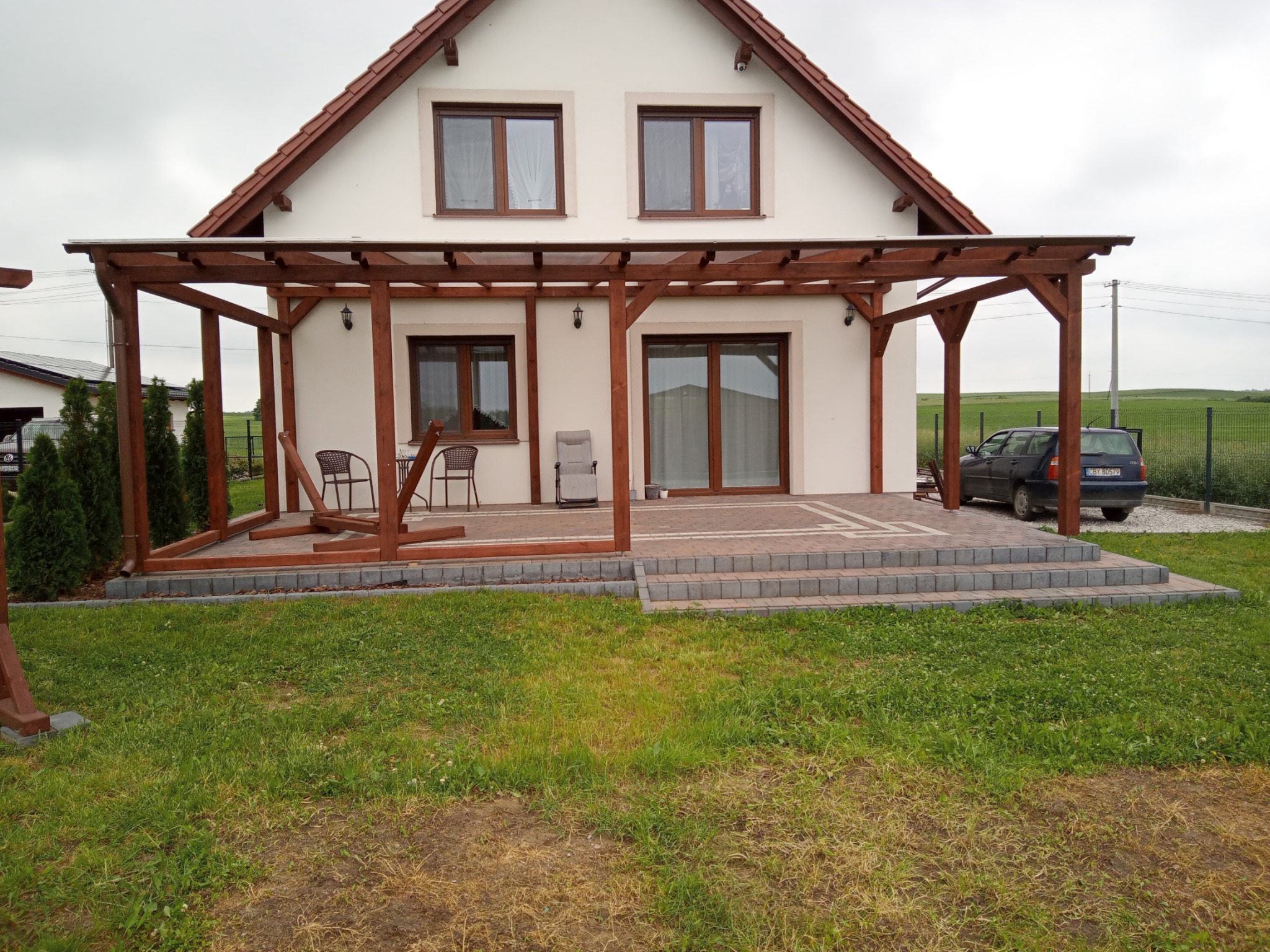 Drewniana konstrukcja pergoli zadaszająca taras przed domem z białymi ścianami i brązowymi oknami, widoczne krzesła ogrodowe i trawnik.