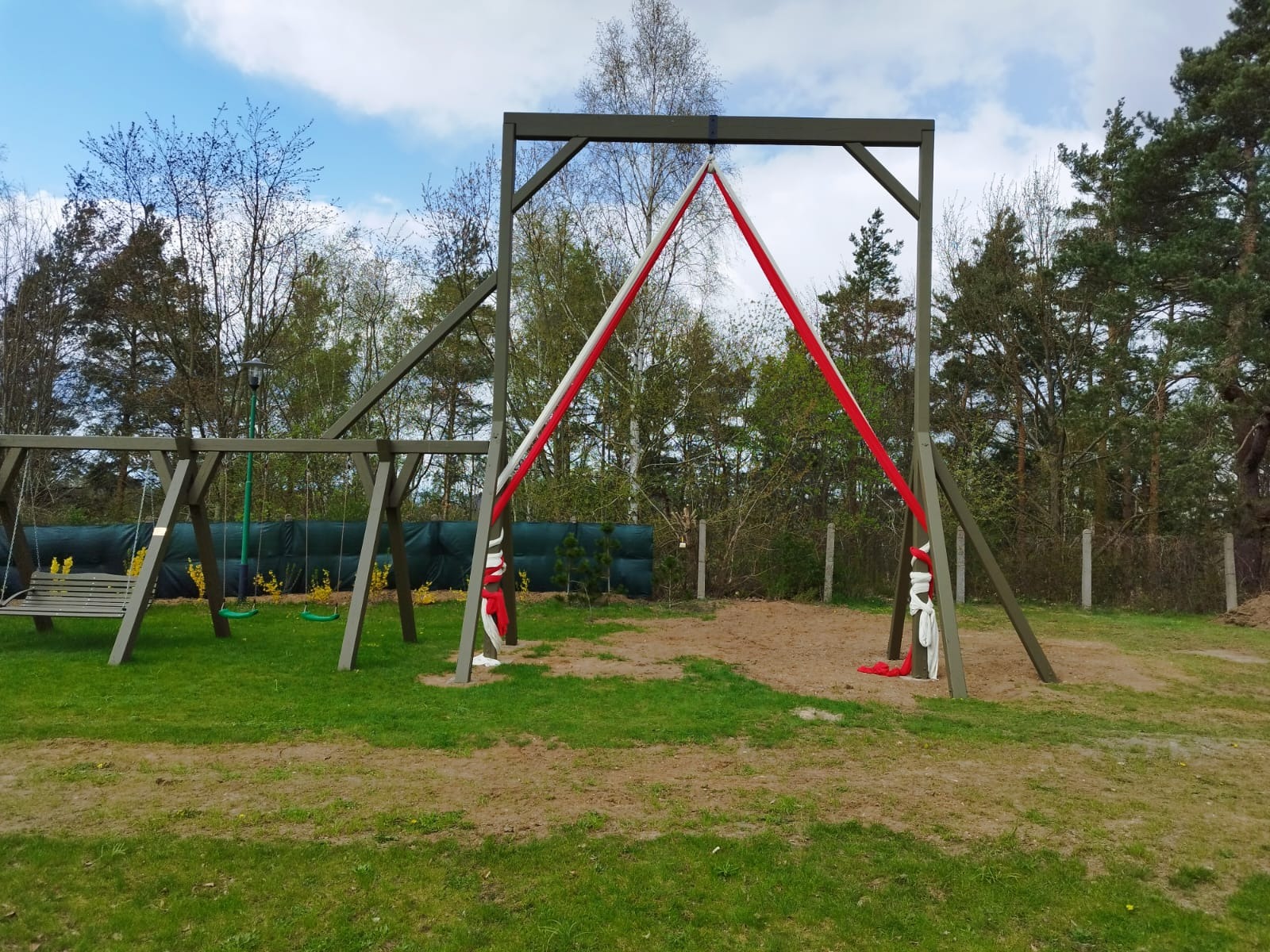 Drewniana konstrukcja huśtawki z zawieszonymi czerwono-białymi wstęgami, stojąca na trawniku z piaszczystym podłożem, w tle las i błękitne niebo.