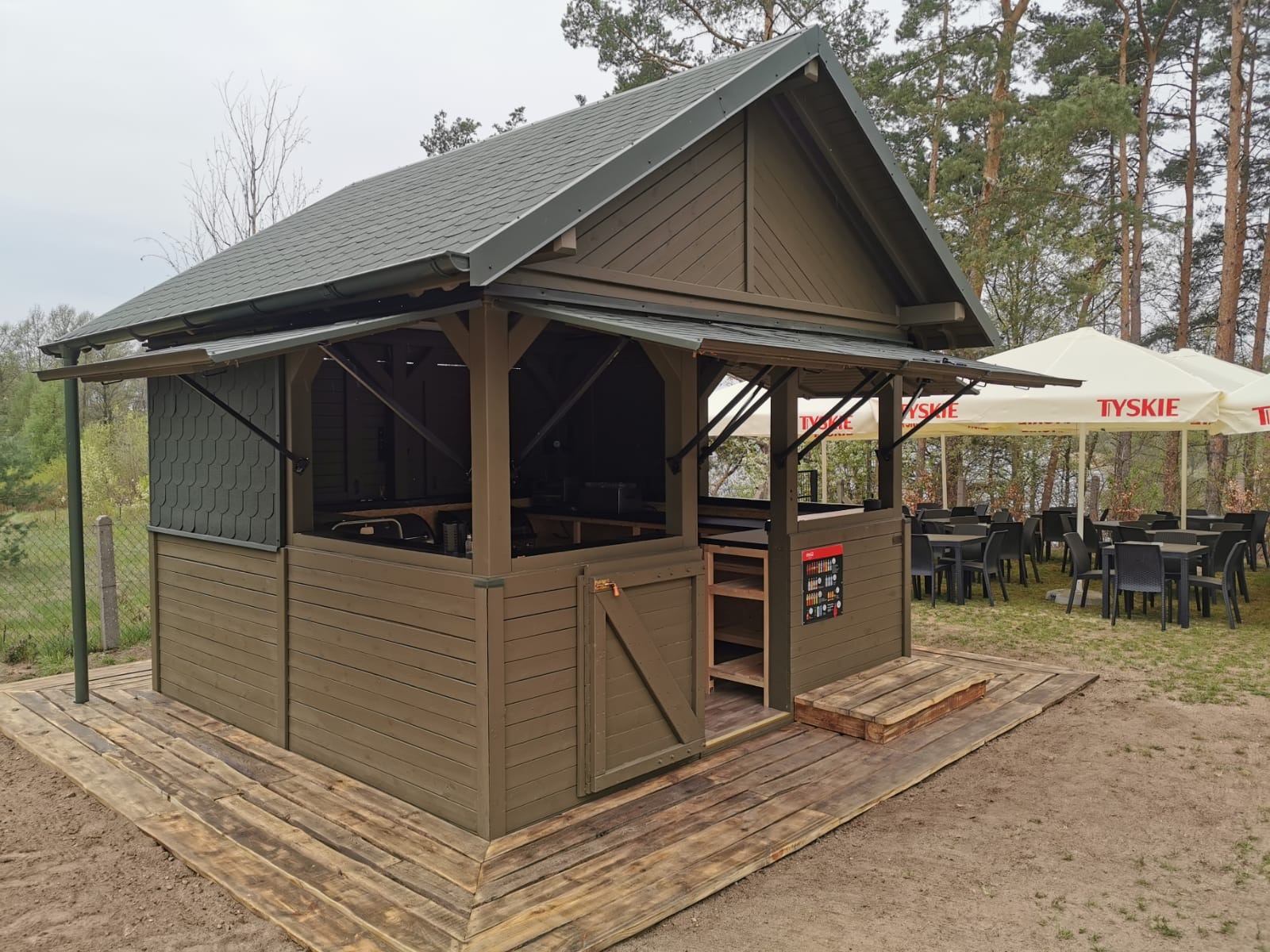 Drewniana altana grillowa w kolorze khaki z zamykanymi okienkami i niewielkim tarasem, widok na stoliki pod parasolami w tle.