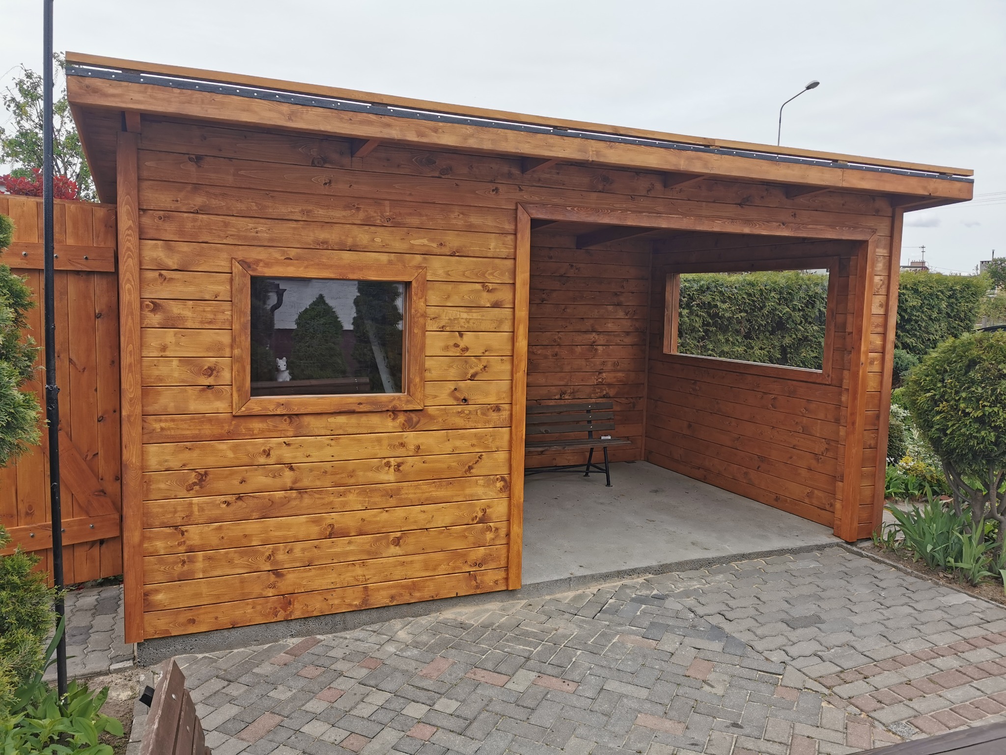 Drewniana altana z oknem, pomalowana na brązowo, z betonową podłogą i ławką w środku, otoczona kostką brukową i zielenią.