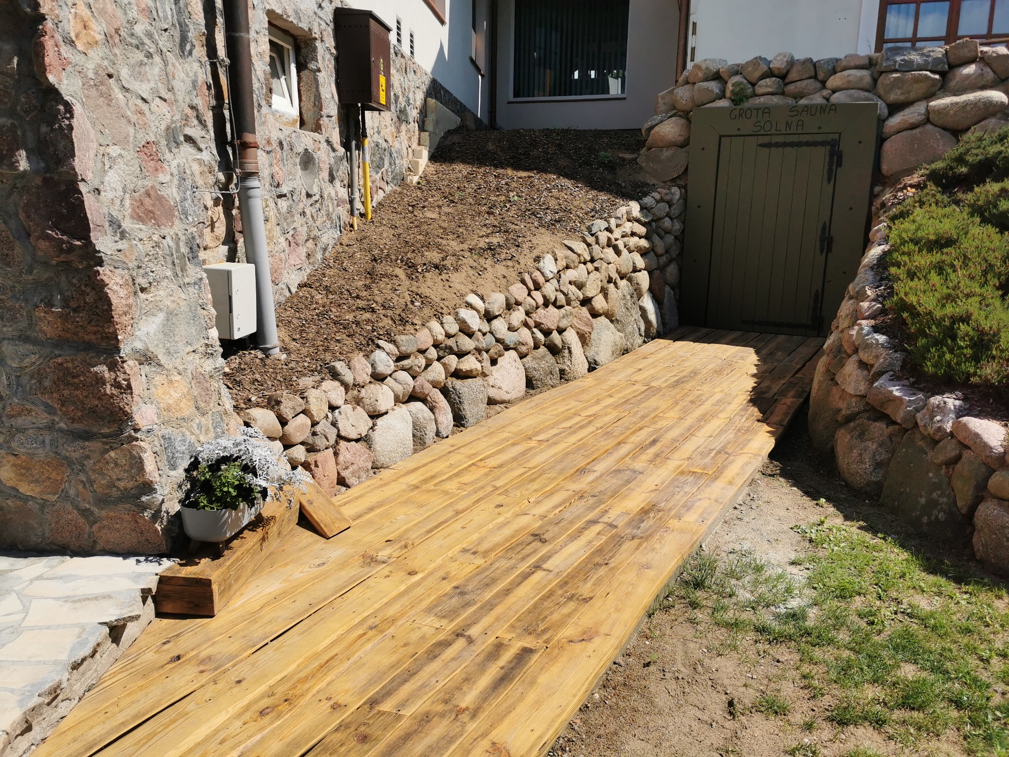 Drewniana ścieżka prowadząca do drzwi z napisem 'Grota Sauna Solna', otoczona kamieniami i roślinnością.