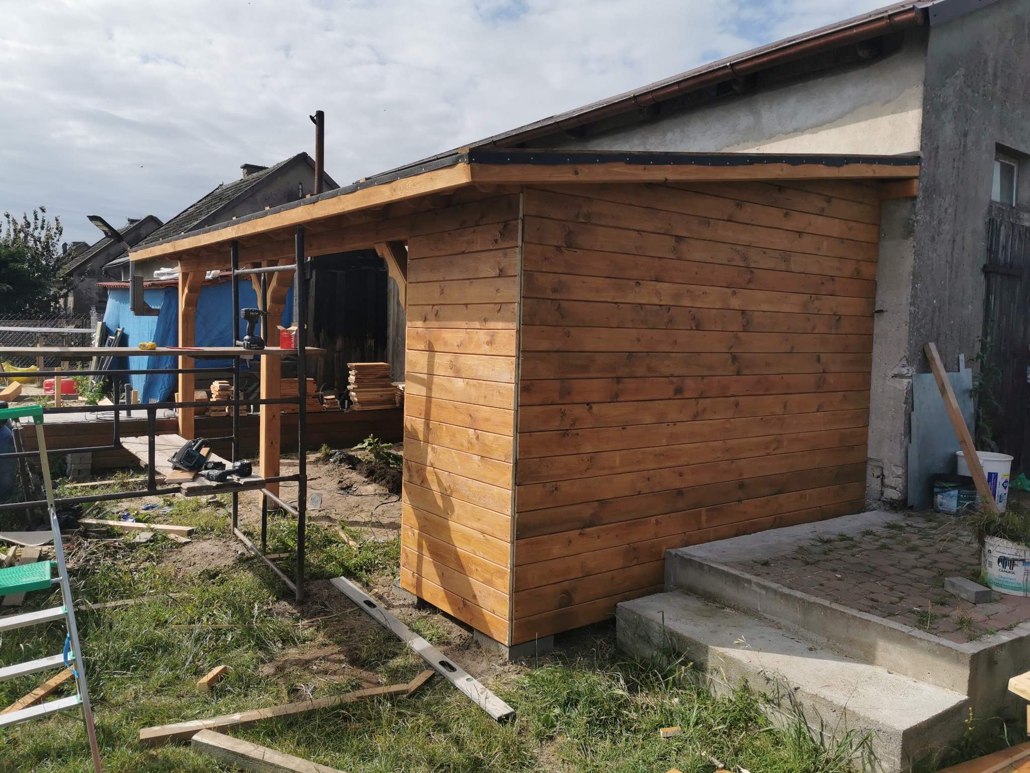 Drewniana wiata w trakcie budowy, widoczne rusztowanie, narzędzia i materiały budowlane, surowe otoczenie z elementami starego budynku.