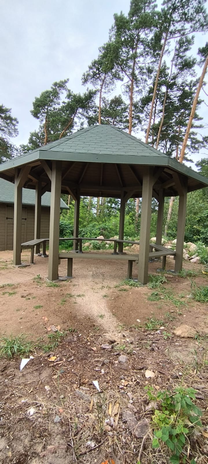 Ośmiokątna, zielona altana z drewnianymi ławkami wewnątrz, otoczona drzewami i zielenią, z widocznymi śladami budowy na ziemi.