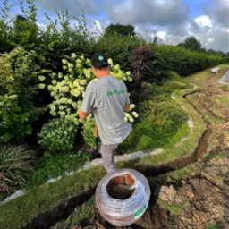 Pracownik w szarej koszulce z logo Rain Bird kopie rów w trawniku obok kwitnących hortensji, przygotowując teren pod instalację systemu nawadniania; zwój brązowego węża w folii leży obok wykopu.