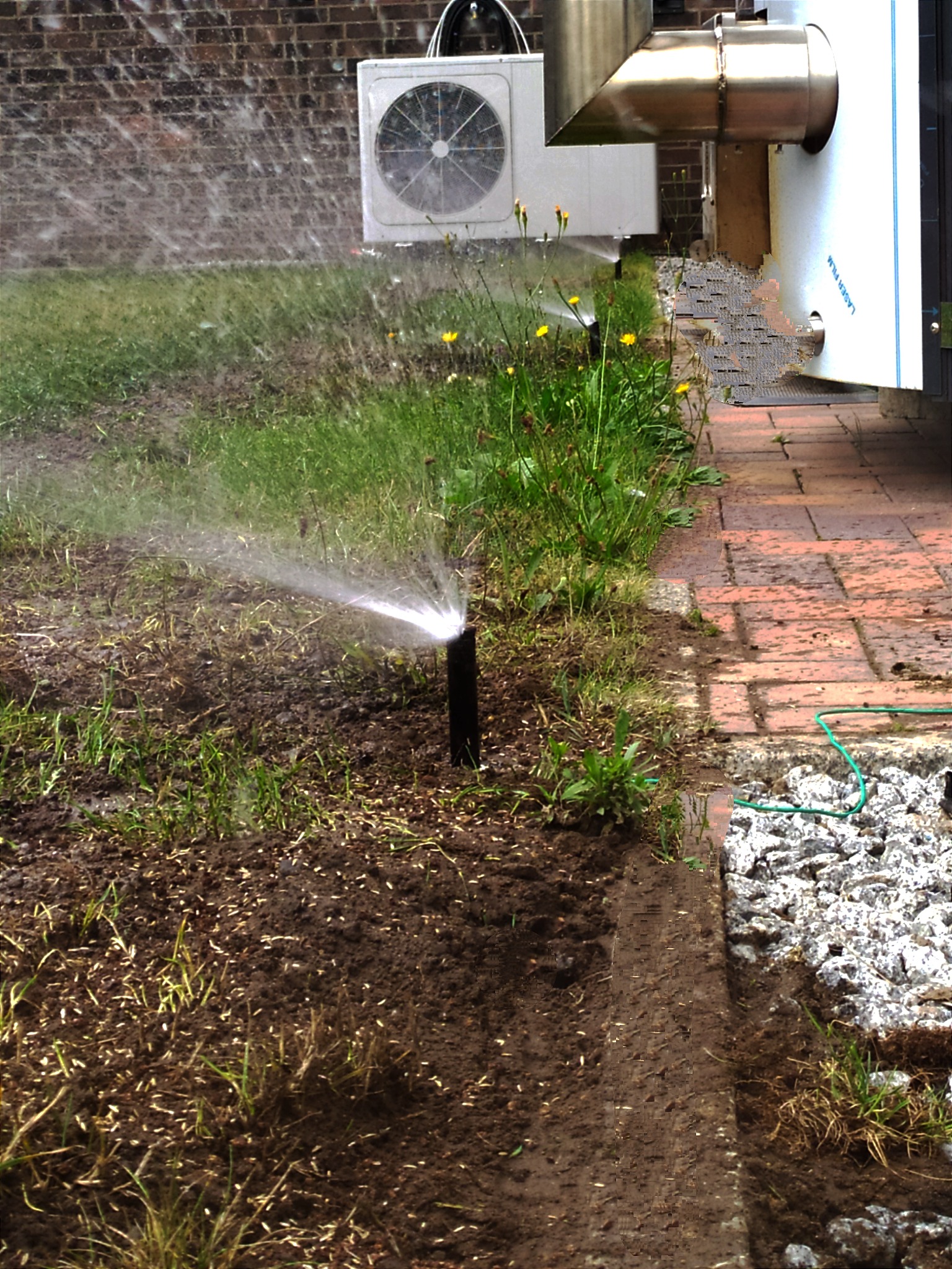 Automatyczny system nawadniania trawnika zraszaczem wynurzalnym, w tle budynek z klimatyzatorem i ceglanym murem.