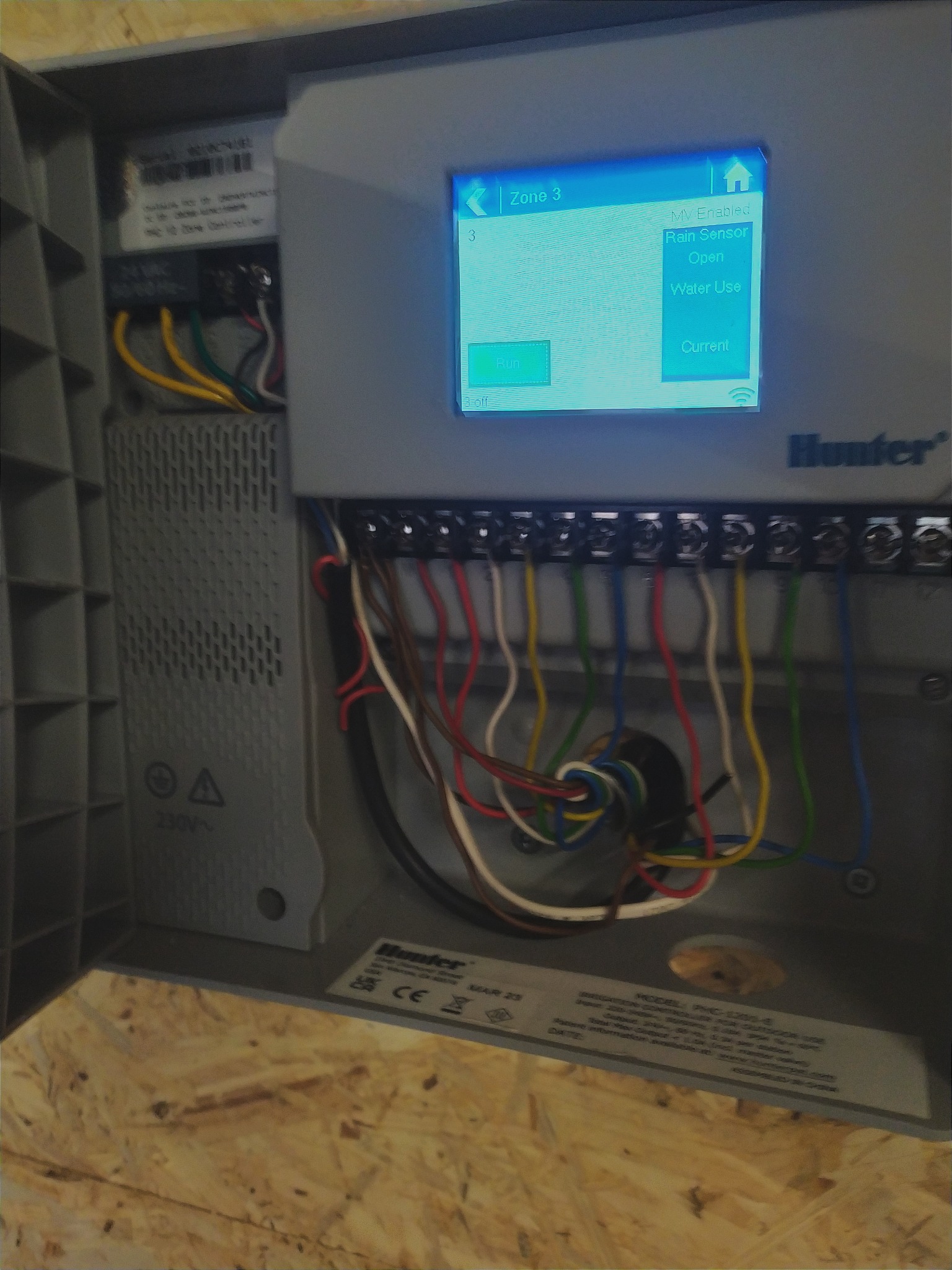 Otwarta skrzynka sterująca systemem nawadniania Hunter, widoczne kolorowe przewody podłączone do listwy zaciskowej oraz ekran LCD z ustawieniami strefy 3, czujnika deszczu i zużycia wody.