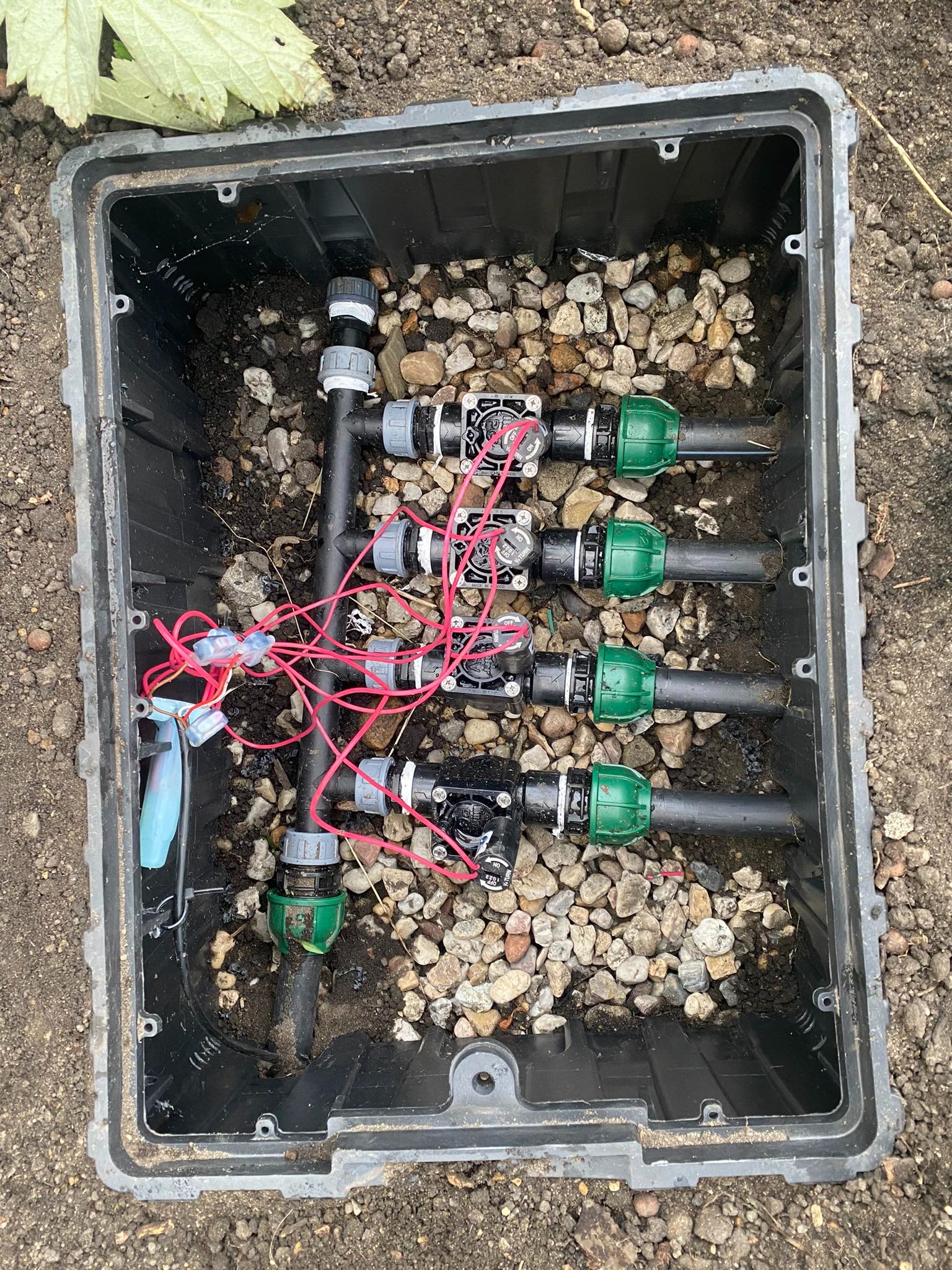 Skrzynka zaworowa z elementami systemu nawadniania, widoczne rury, zawory elektromagnetyczne i okablowanie, wypełniona drobnymi kamieniami dla ochrony przed zamarzaniem.