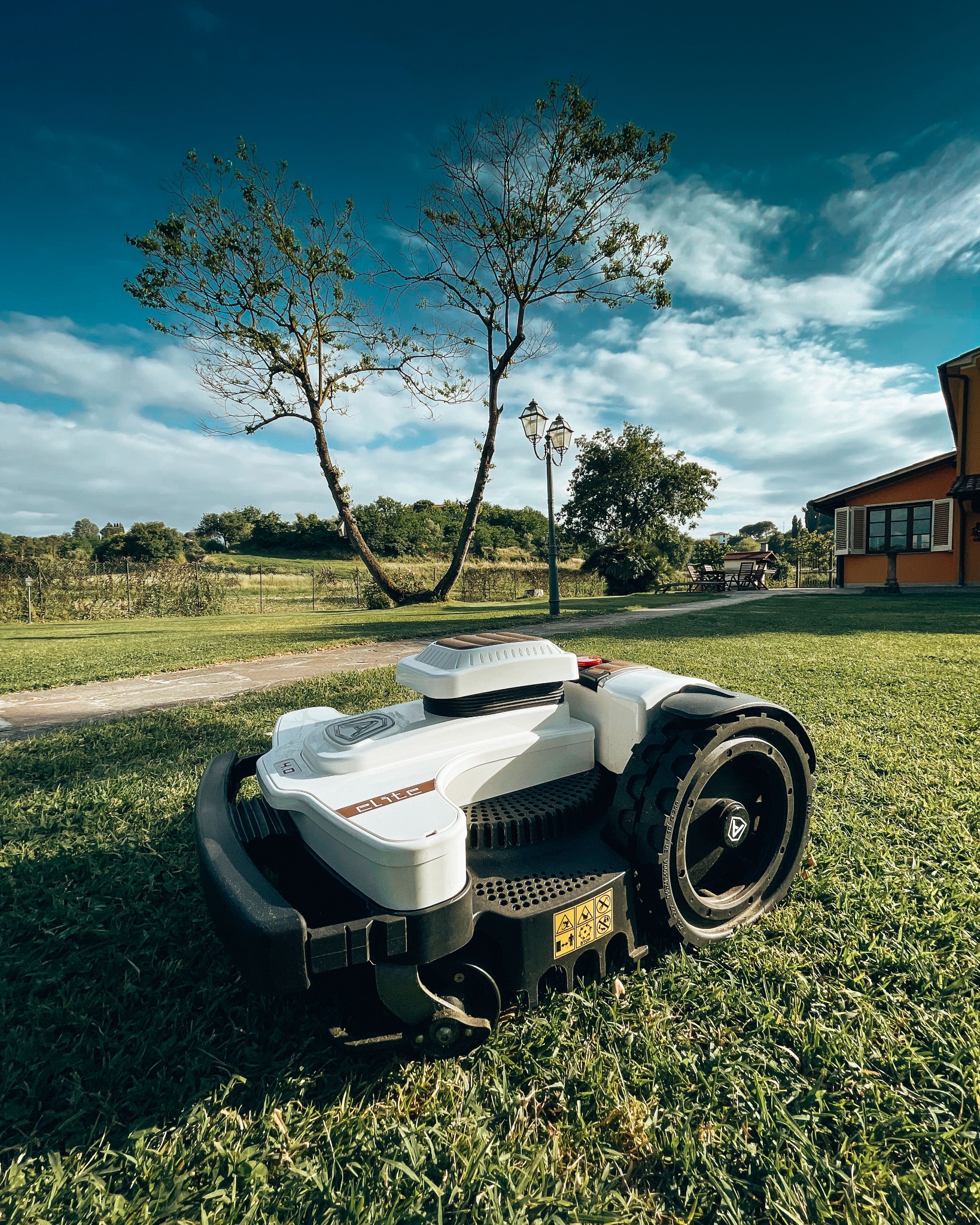 Automatyczna kosiarka do trawy, model Elite, na zadbanym trawniku z widokiem na drzewa i budynek w tle, błękitne niebo z chmurami.