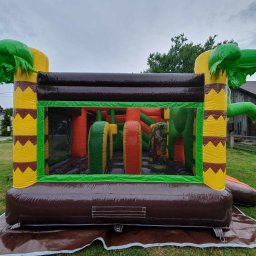 PSotnik Dmuchańce Patrycja Szczybyło - Kolorowy dmuchany plac zabaw w stylu dżungli z motywem dinozaurów, ustawiony na trawie. Widoczne elementy dekoracyjne imitujące drzewa i przeszkody wewnątrz.