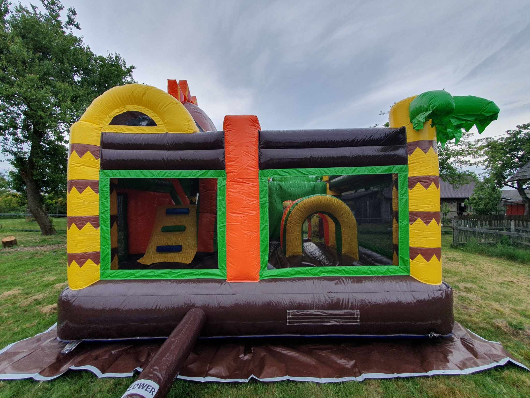 Kolorowy, dmuchany plac zabaw w stylu dinozaurów na trawie, z tunelem i ścianką wspinaczkową. Widoczne detale: imitacja palm, brązowa podstawa, żółte elementy dekoracyjne.