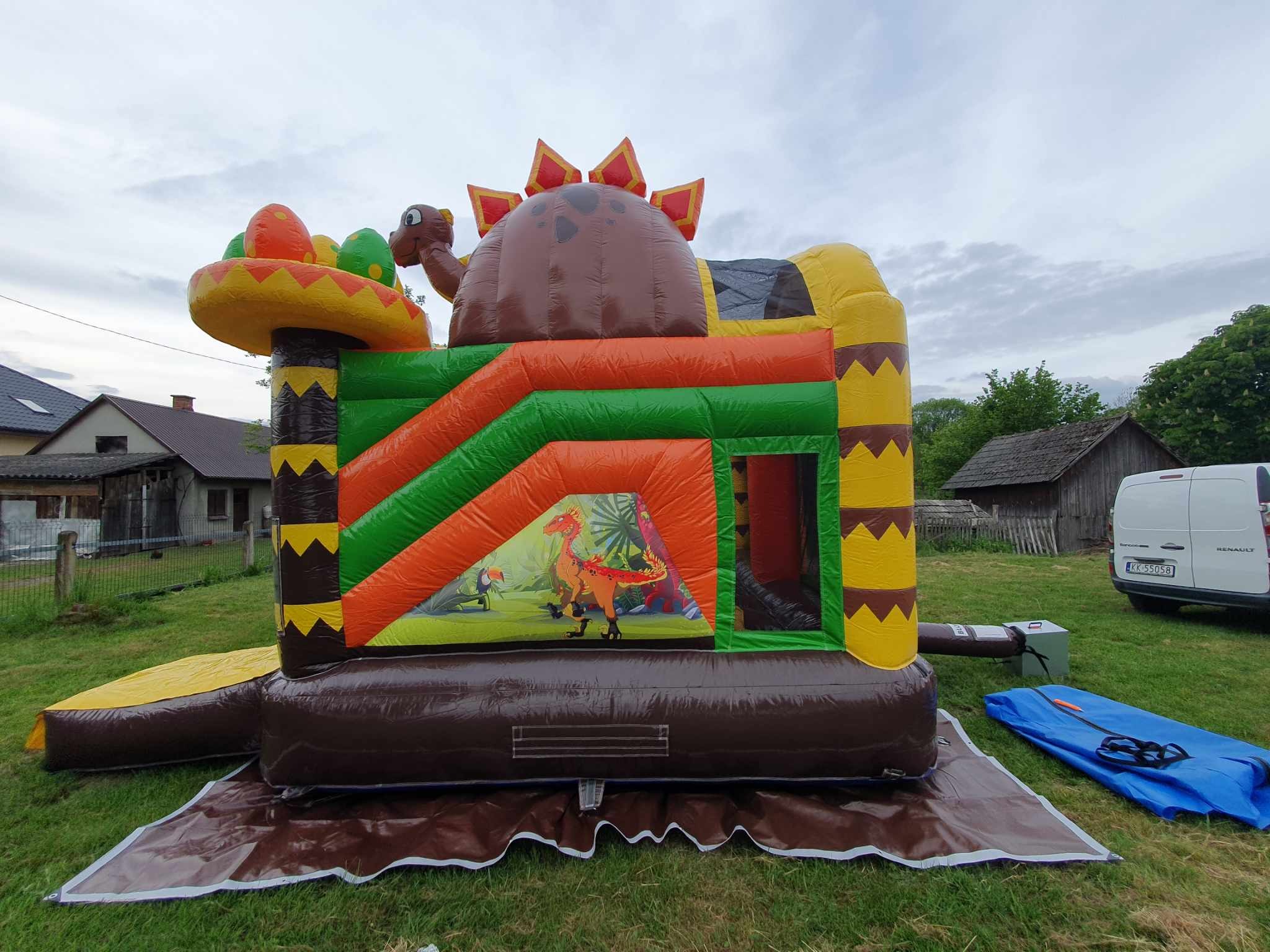 Kolorowy dmuchany plac zabaw w kształcie dinozaura z motywami dżungli, ustawiony na trawie przed budynkiem mieszkalnym i zaparkowanym białym vanem.