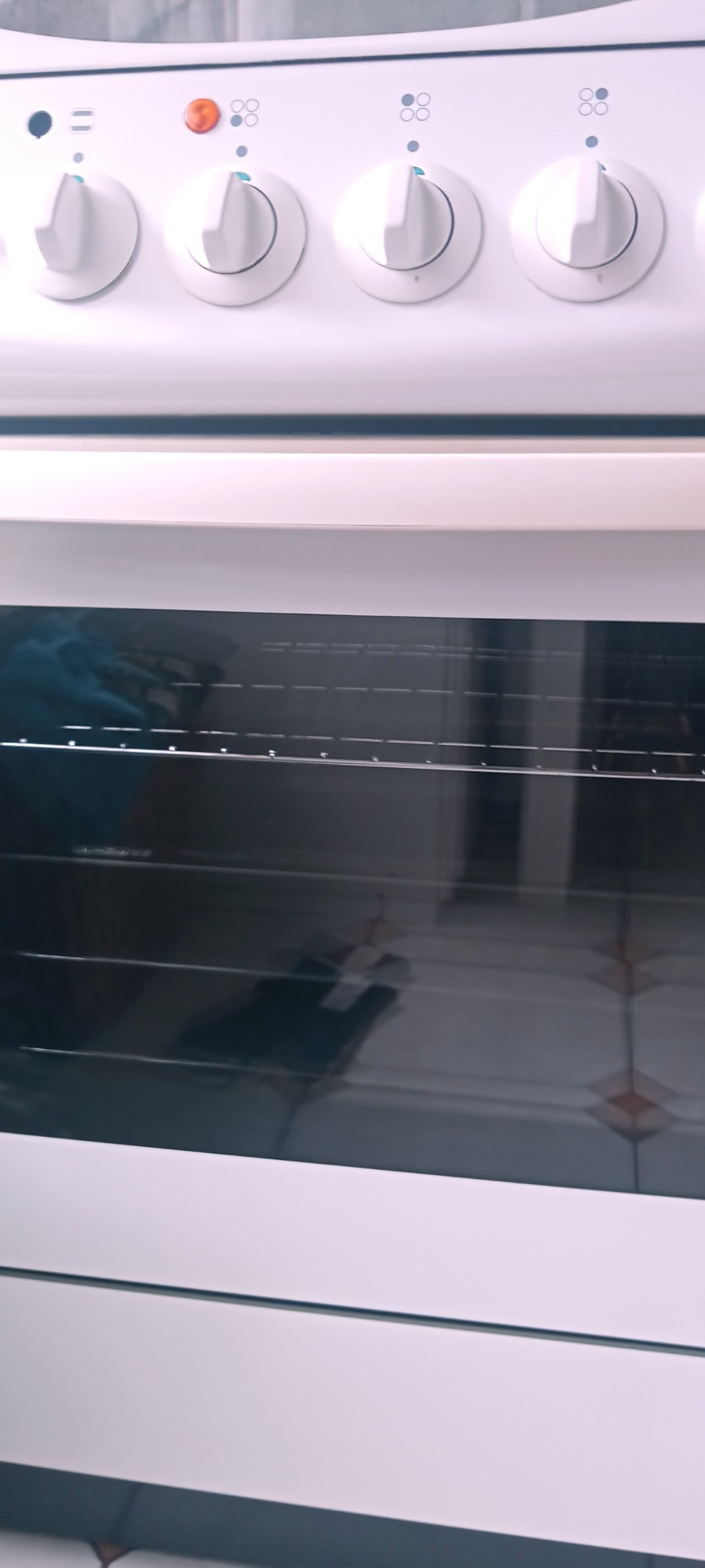 Biała kuchenka z widocznym wnętrzem piekarnika, na dnie którego leżą ciemne przedmioty, a na górze rząd białych pokręteł.