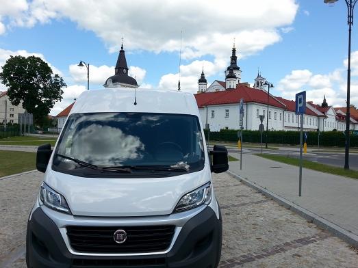 Biały bus Fiat zaparkowany na brukowanej ulicy, z odbiciem chmur na przedniej szybie, na tle klasztoru z wieżami i znaków drogowych.