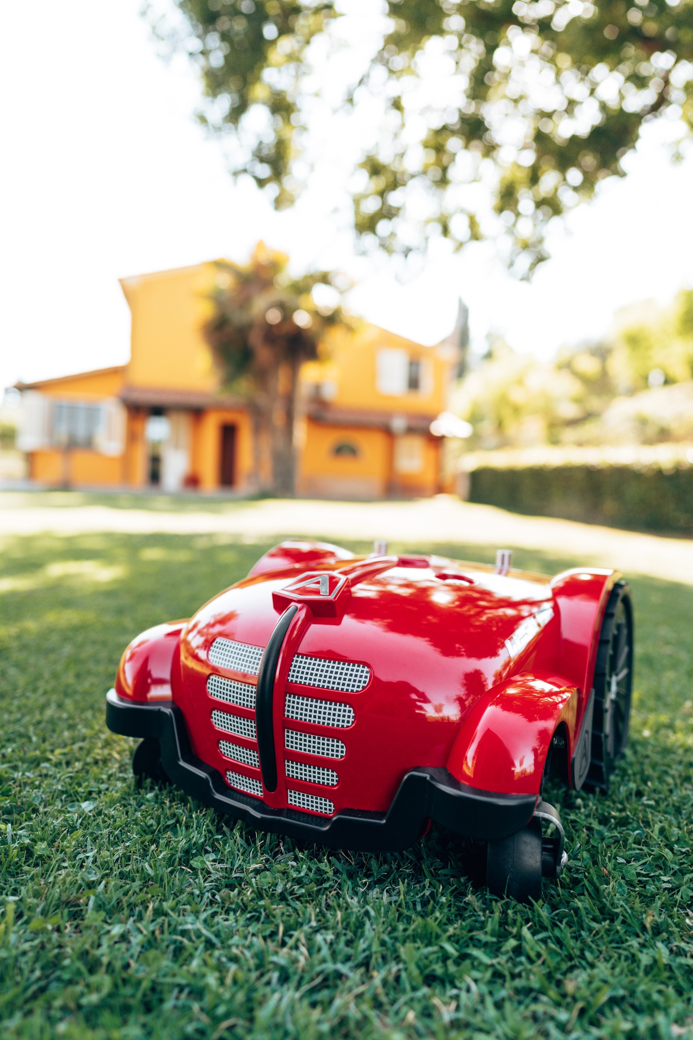 Czerwony, autonomiczny robot koszący na trawniku przed żółtym domem w tle, z widocznym logo firmy na obudowie.
