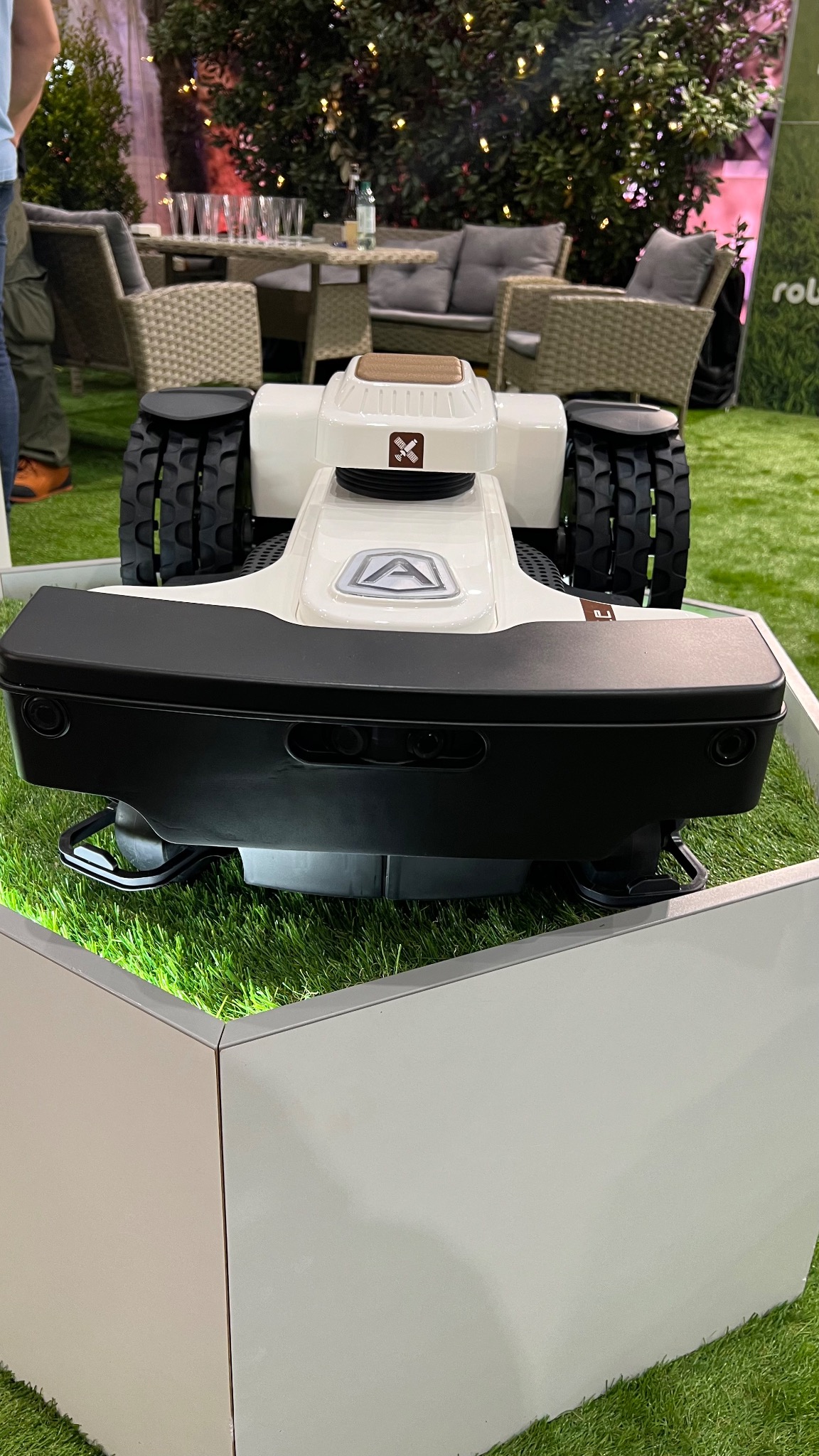 Robot koszący trawnik z gumowymi kołami, prezentowany na sztucznej trawie wewnątrz białej obudowy, w tle meble ogrodowe i dekoracyjne rośliny.