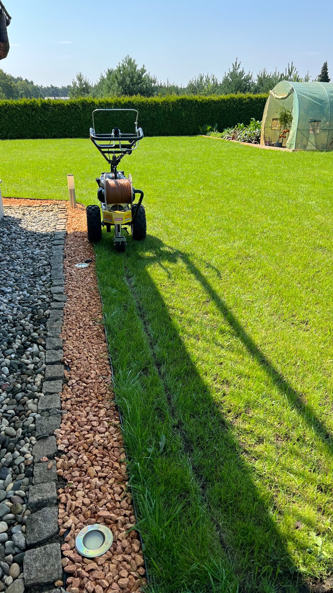 Profesjonalny aerator do trawy tworzy równą linię w trawniku, obok krawężnika z kamieni ozdobnych i lampą gruntową, w tle żywopłot i szklarnia.