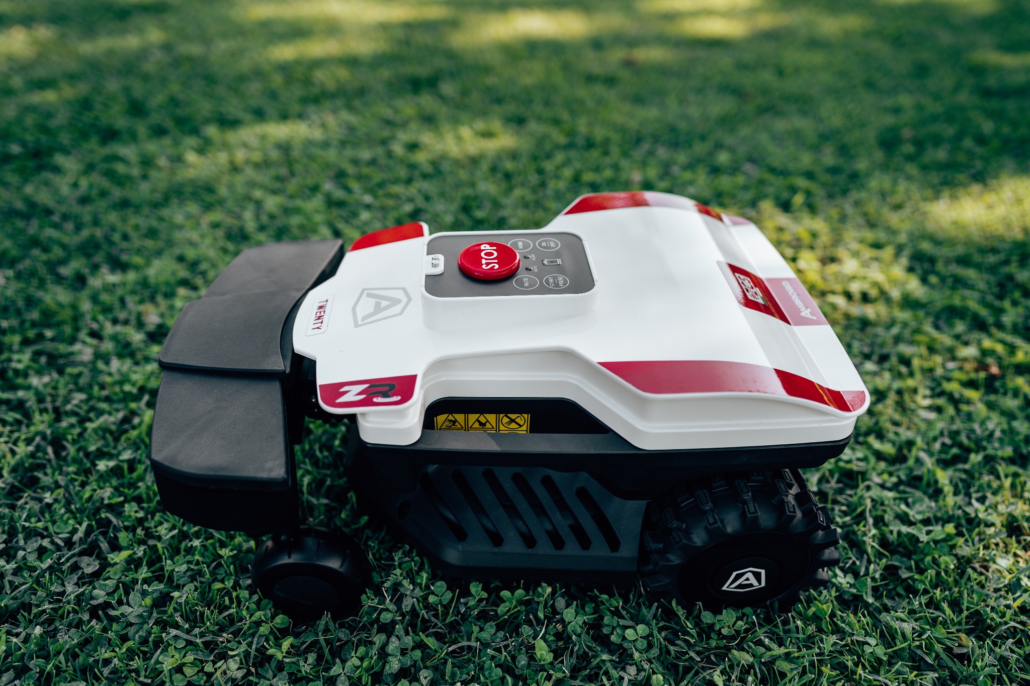 Automatyczna kosiarka do trawy z czerwonym przyciskiem STOP na panelu sterowania, stojąca na krótkiej, zielonej trawie z koniczyną.
