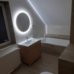 Nowoczesna łazienka na poddaszu z okrągłym lustrem podświetlanym LED, drewnopodobne płytki, biała armatura i skośny sufit.