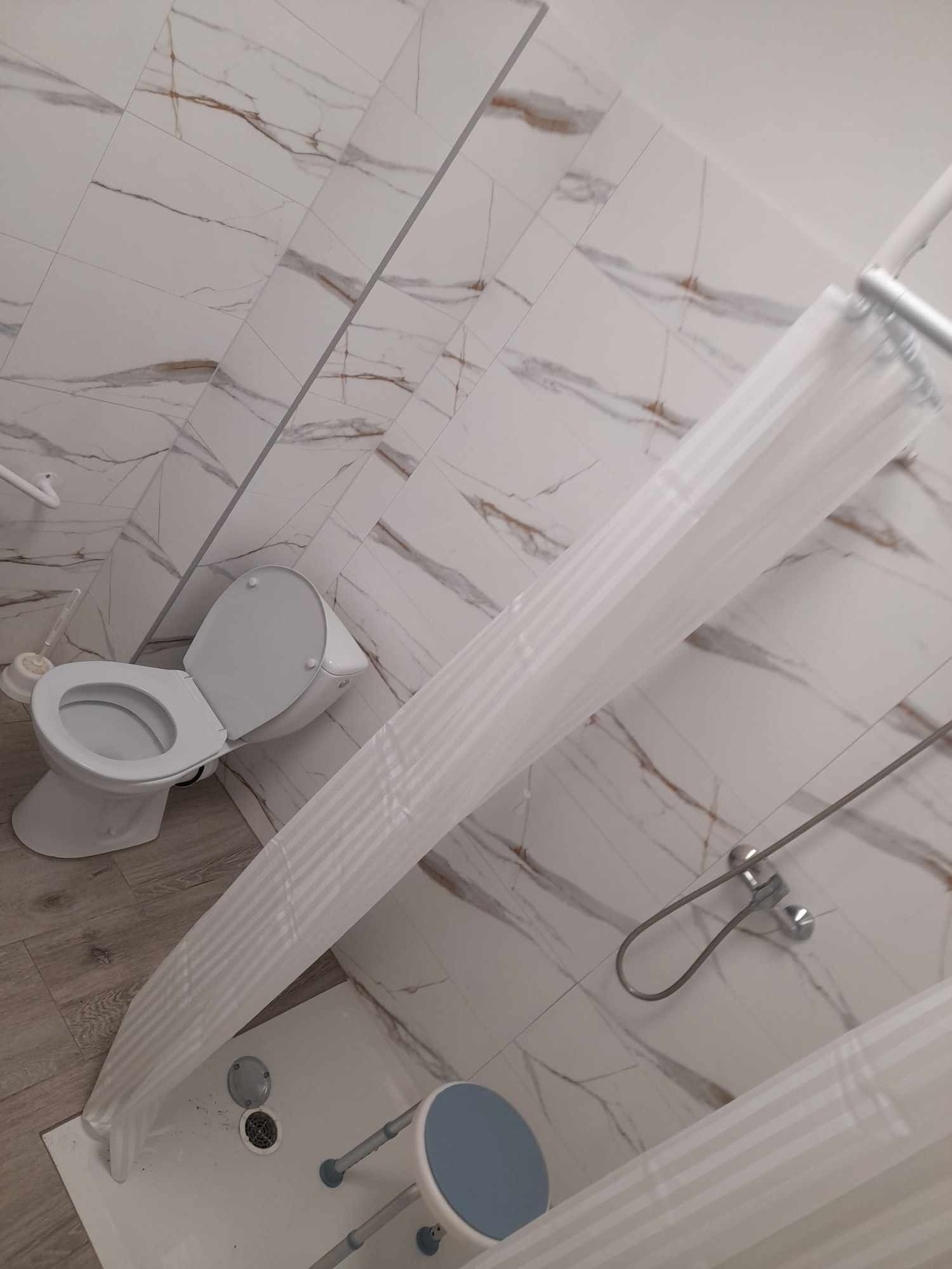 Jasna łazienka po remoncie z marmurowymi płytkami na ścianach, widoczna toaleta z otwartą klapą, prysznic z zasłoną i stołkiem dla osoby niepełnosprawnej.