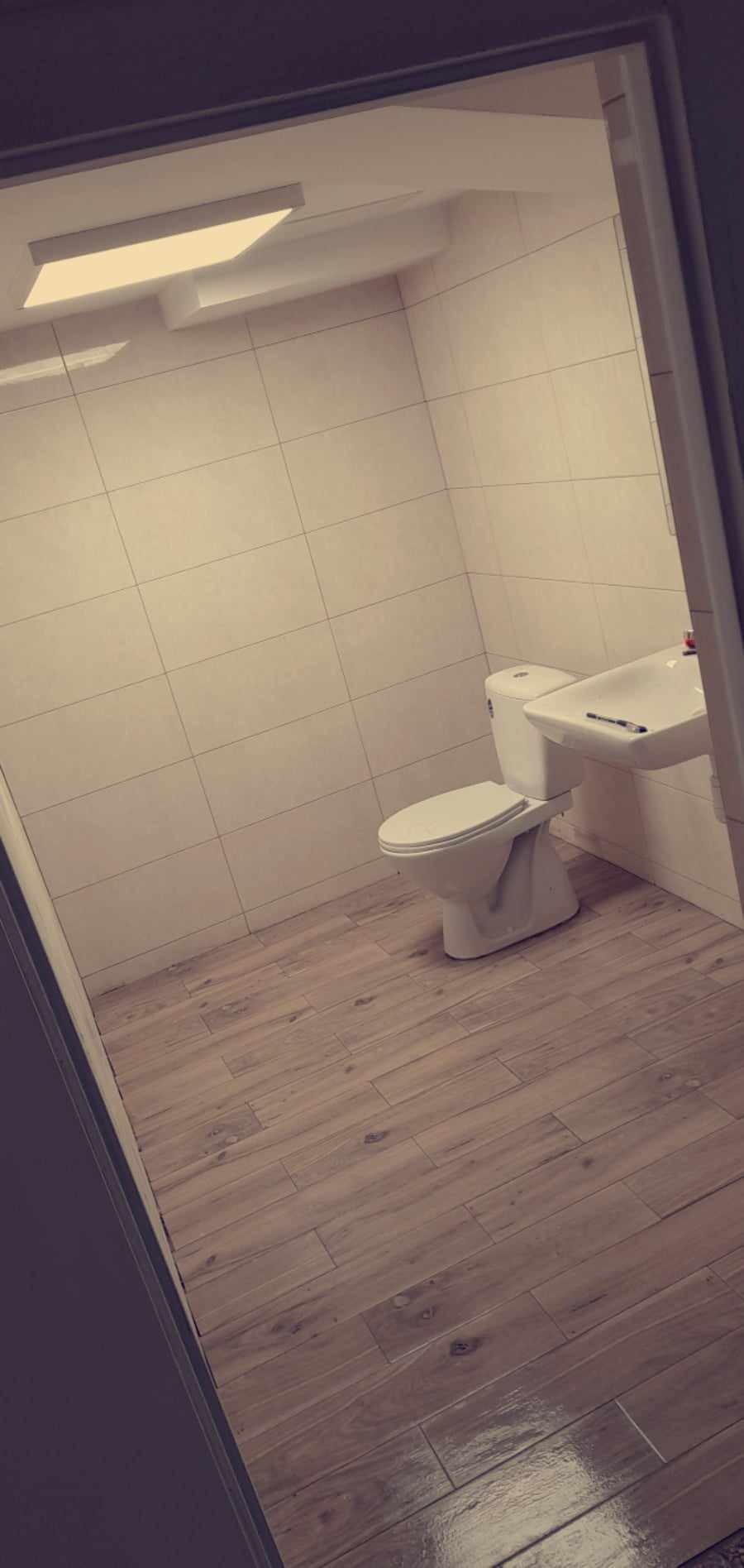 Wykończona łazienka z beżowymi płytkami na ścianach i podłodze imitującymi drewno, widok na toaletę i umywalkę z bliska, ujęcie zza framugi drzwi.