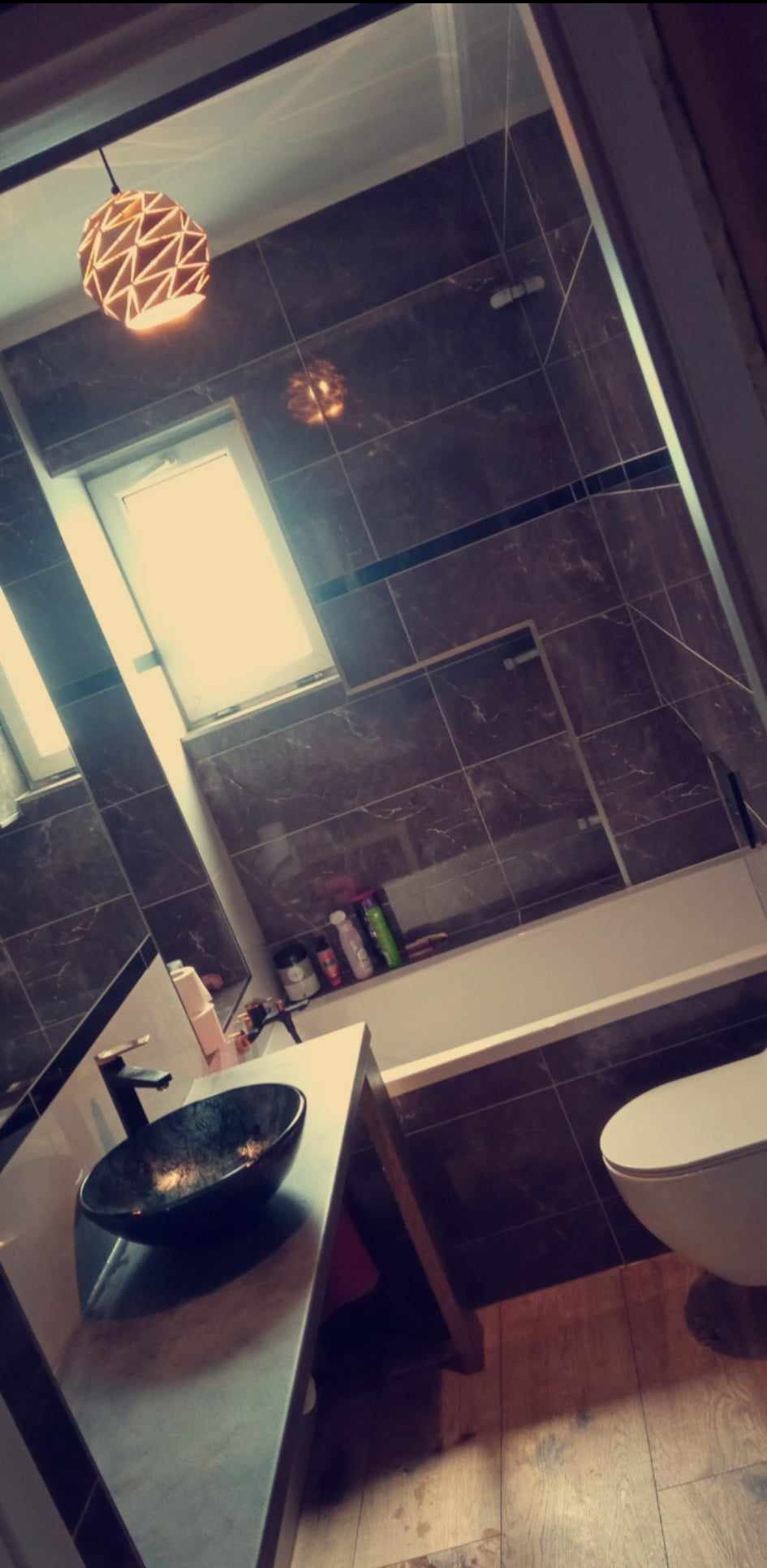 Nowoczesna łazienka z brązowymi, marmurkowymi płytkami, owalną umywalką w kolorze czarnym i lampą z geometrycznym wzorem, widziana pod kątem.