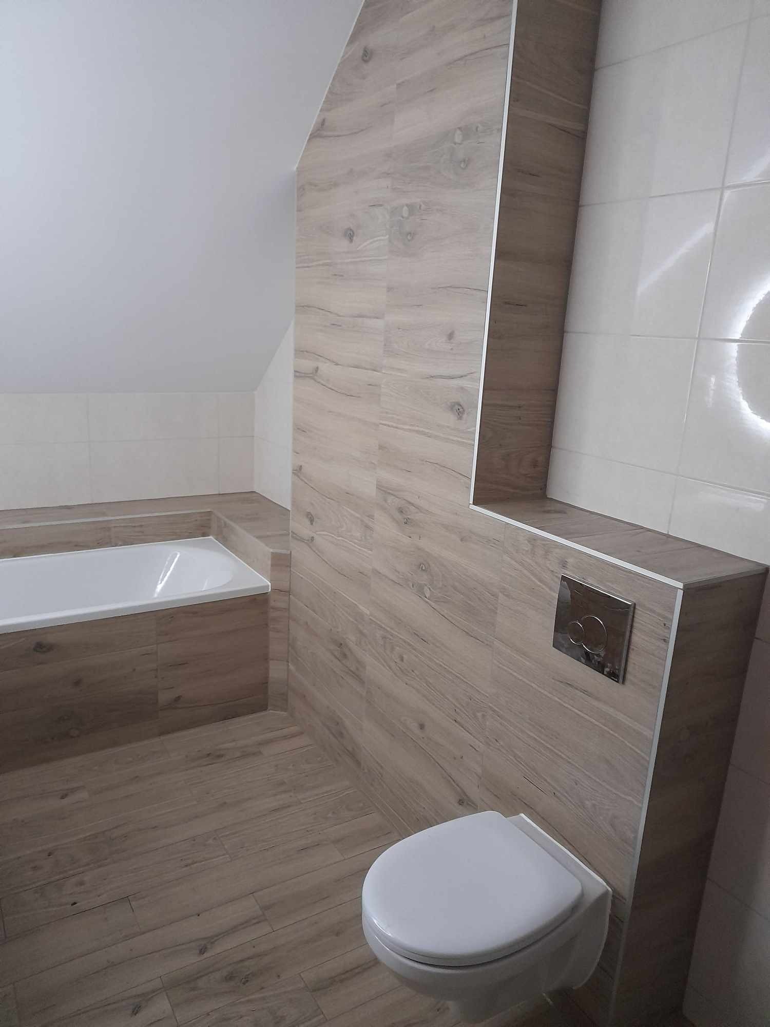 Wykończona łazienka na poddaszu z wanną, toaletą podwieszaną i płytkami imitującymi drewno na ścianach i podłodze, z widocznym przyciskiem spłukującym.