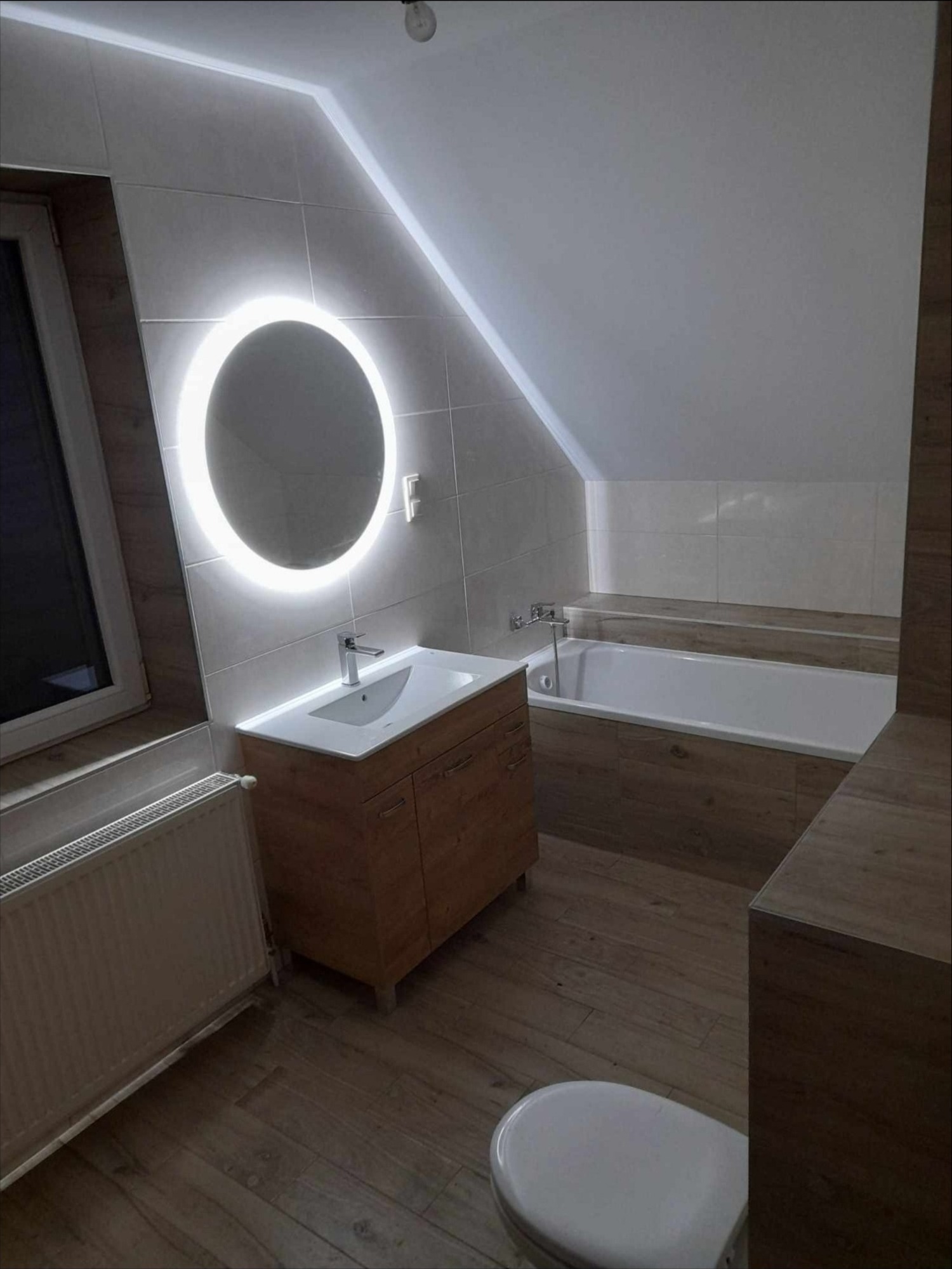 Nowoczesna łazienka na poddaszu z okrągłym lustrem podświetlanym LED, drewnopodobne płytki, biała armatura i skośny sufit.